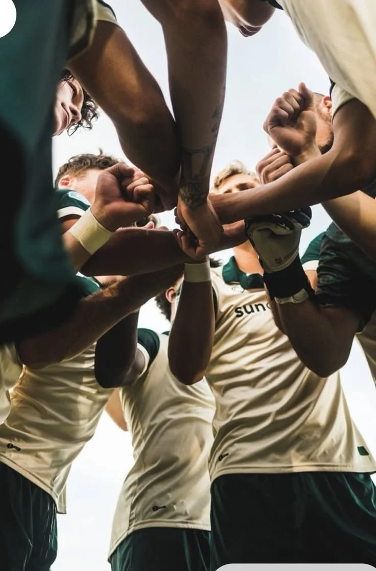 A group of soccer players in a huddle with their hands stacked together before a game or practice. They are wearing sports gear and are outdoors.