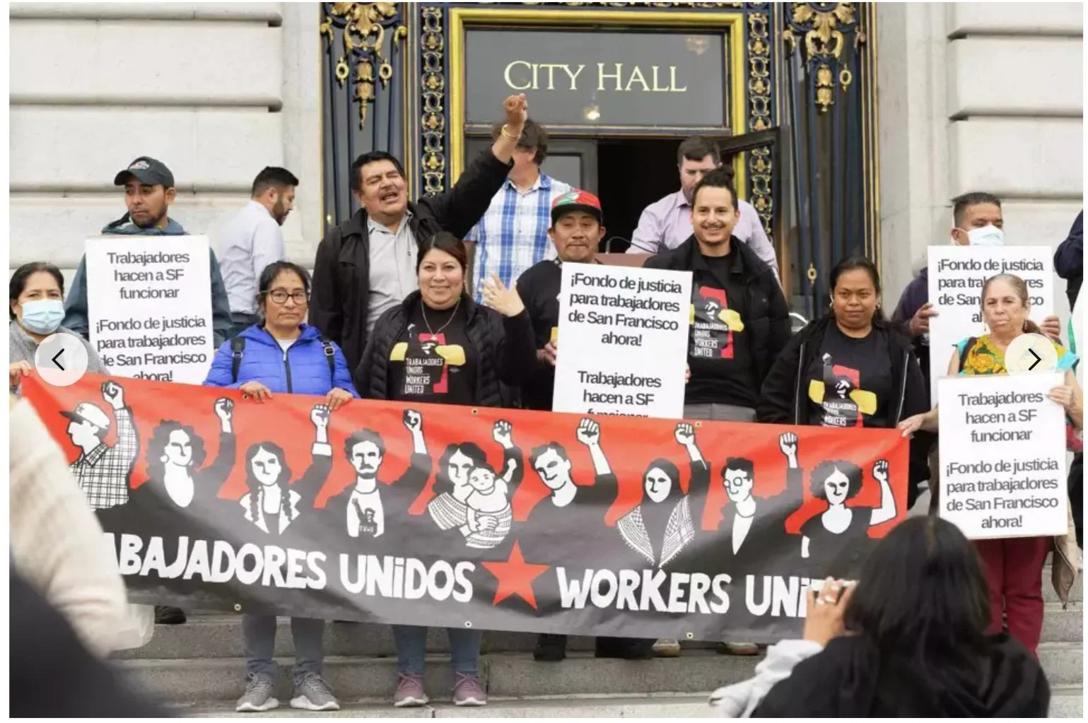 A group of demonstrators stand in front of City Hall holding signs and a banner.
