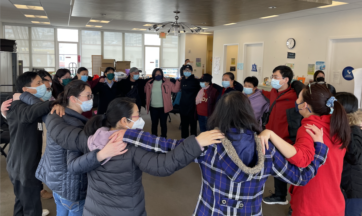 Approximately 20 people stand in a circle, holding each other by the shoulders.