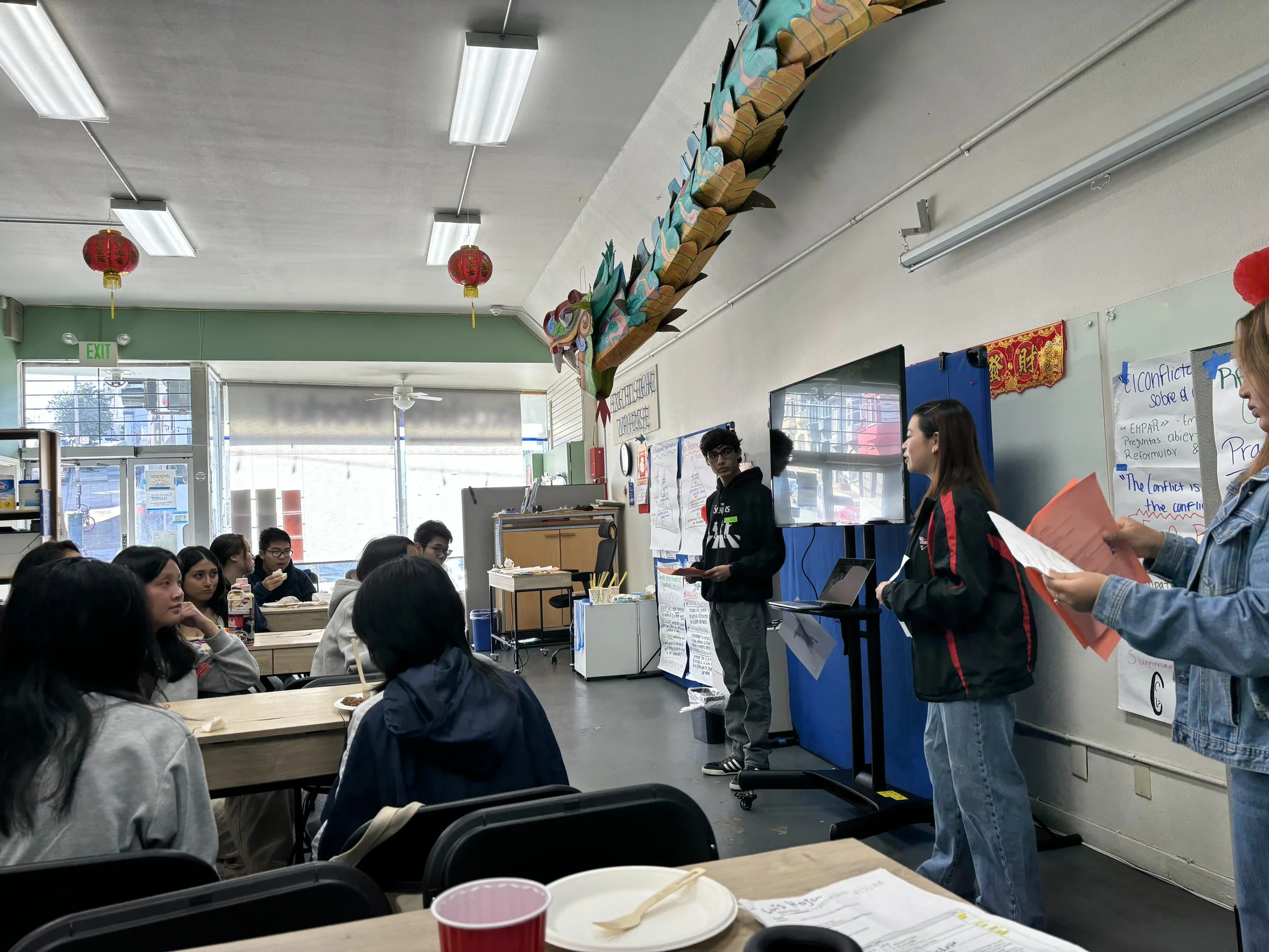 Two youth stand at the side of the room. about 10-15 youth sitting and listening to the presentation.