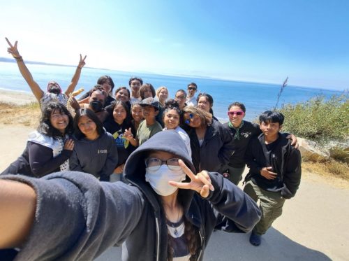 A selfie of about 20 young people. There is a large body of water in the background.
