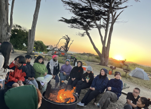 13 people sit around a campfire. There is a tent and some trees in the background. The sun is setting in the distance.