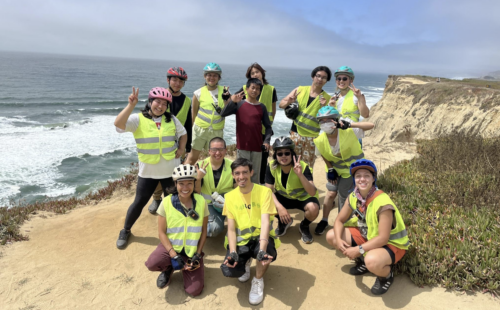 A group photo of 13 young people standing and sitting on a beach with water in the background. All but one of them are wearing reflective vests, and some are wearing bike helmets.