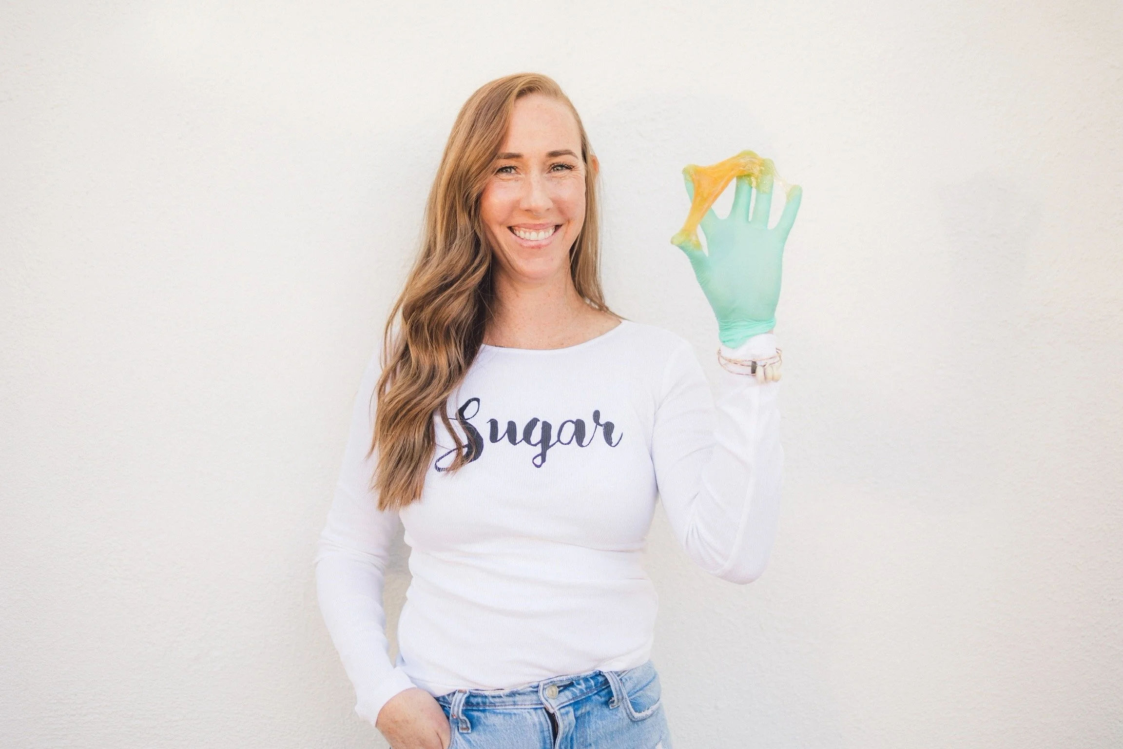 A woman with long wavy hair smiling and holding a green cleaning glove with yellow sponge on her hand, wearing a white shirt with 'Sugar' written on it and blue jeans, standing against a plain light-colored wall.