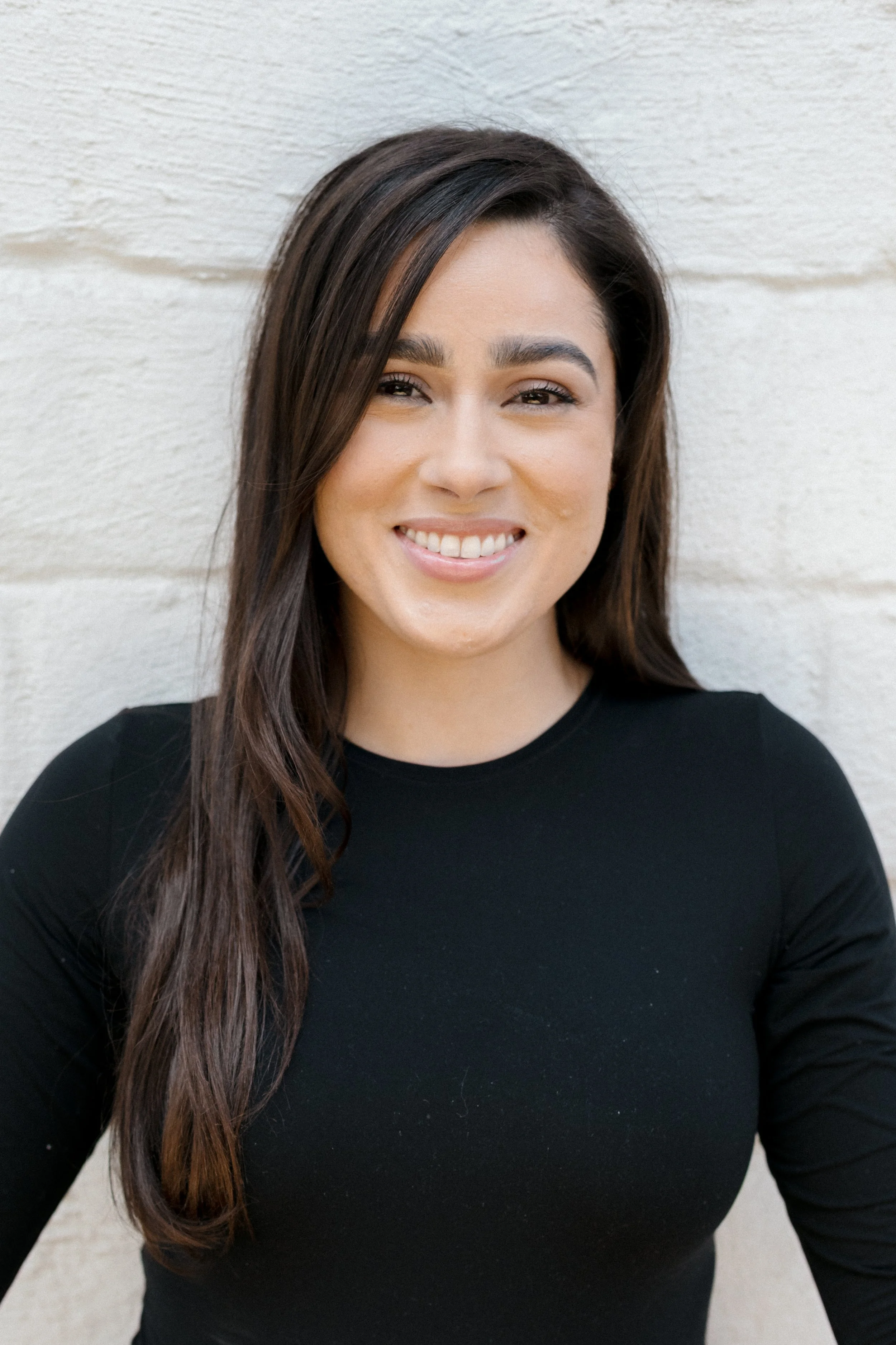 Young woman with curly hair, smiling, wearing a white shirt with the word "Sugar" and holding a gummy bear in her hand, standing against a plain wall.