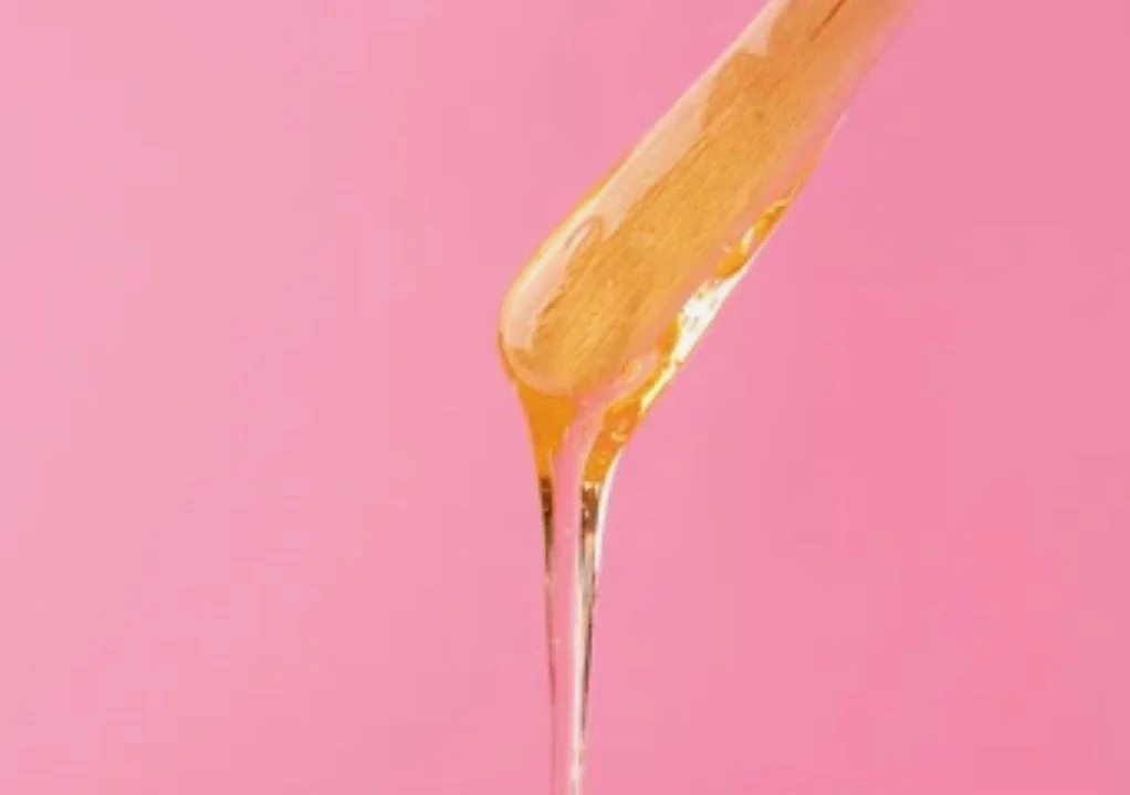 Honey being drizzled from a wooden stick onto a pink background.