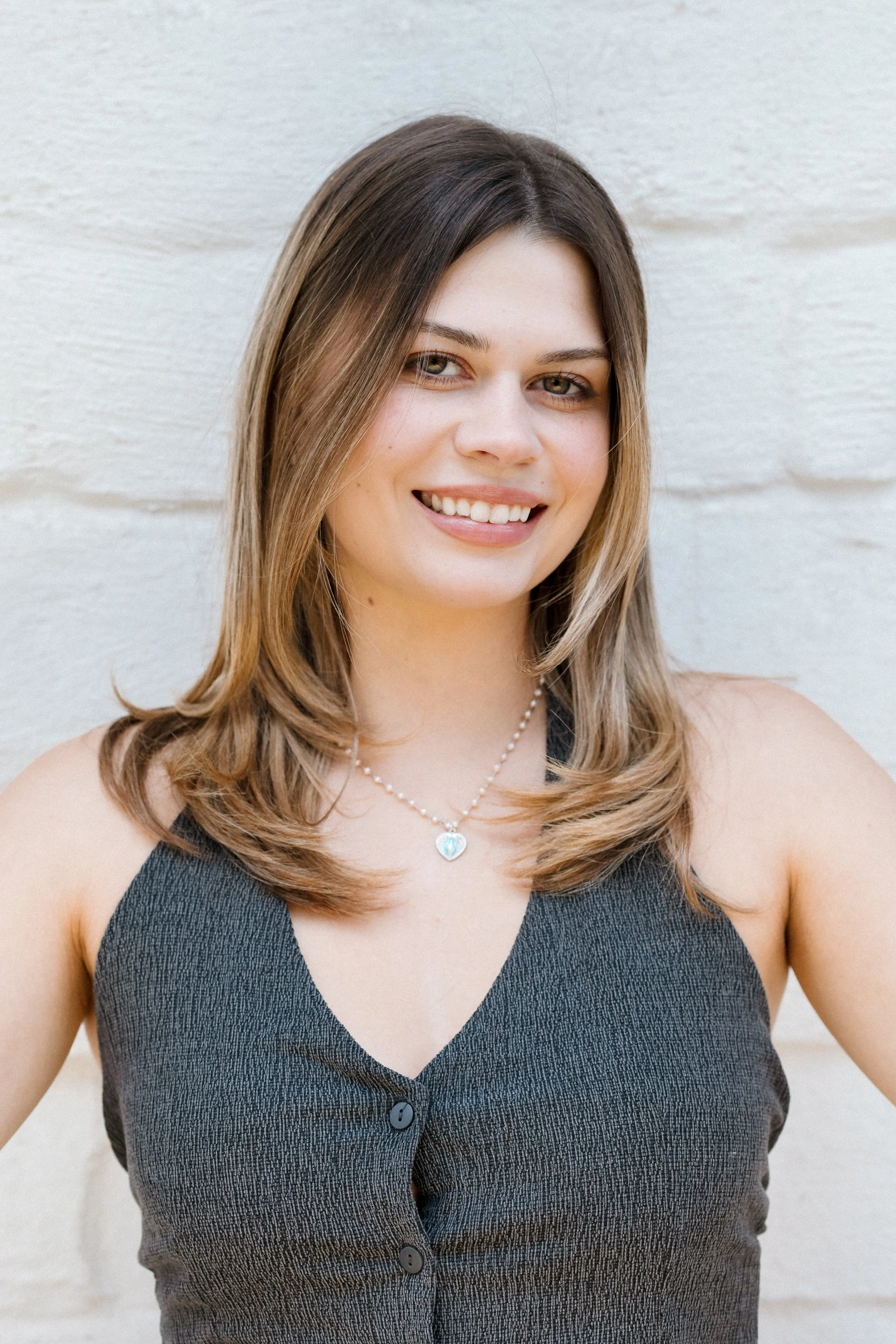A woman with shoulder-length brown hair wearing a cream-colored sweater with the word "Sugar" on it, blue jeans, and a necklace with an "S" pendant, smiling and holding a yellow gummy candy in her gloved hand against a white textured wall.