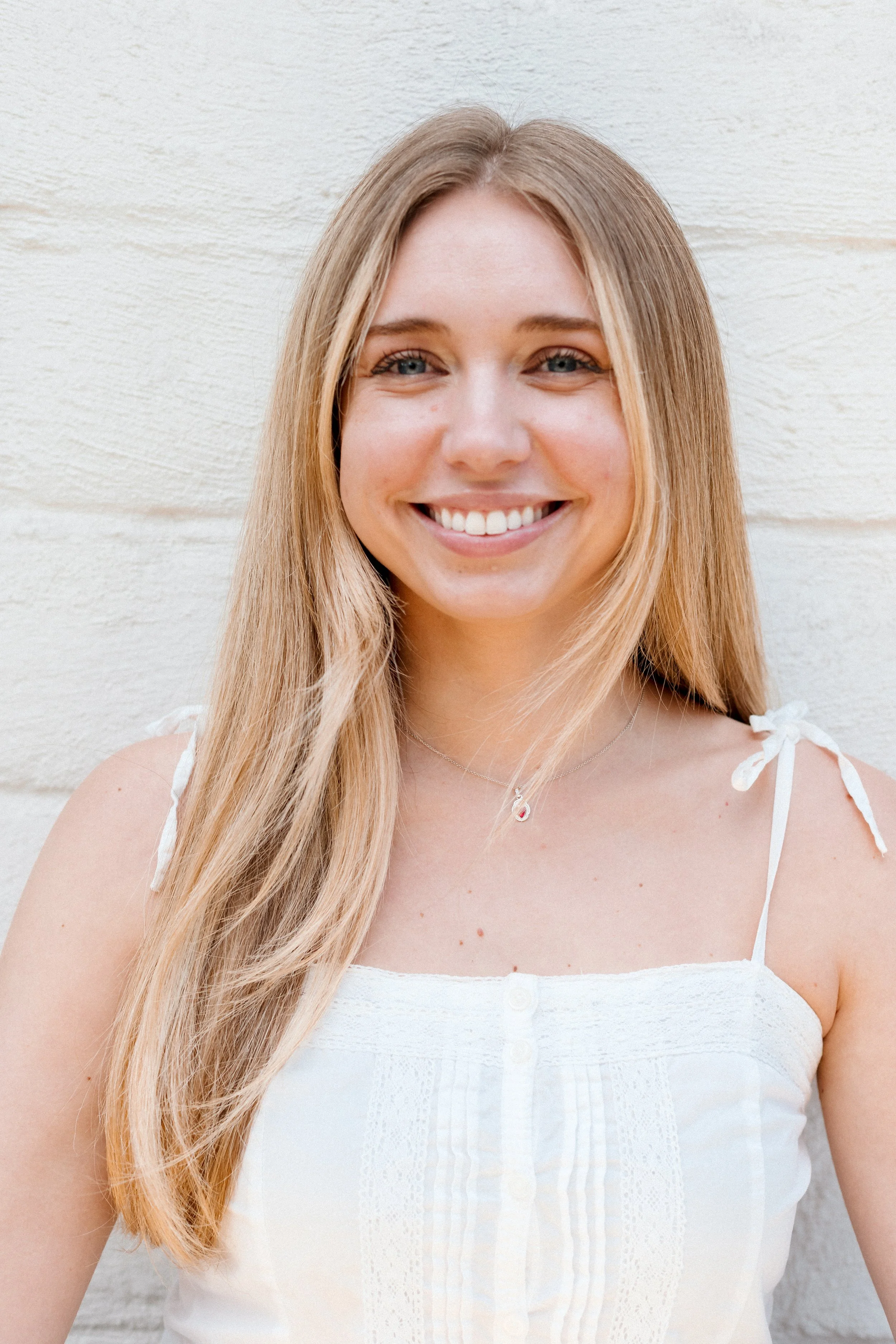 A young woman with long, wavy blonde hair smiling, wearing a white long-sleeve shirt with black text that says 'Sugar', and a green glove on her right hand, holding a yellowish gummy candy against a plain light-colored wall.