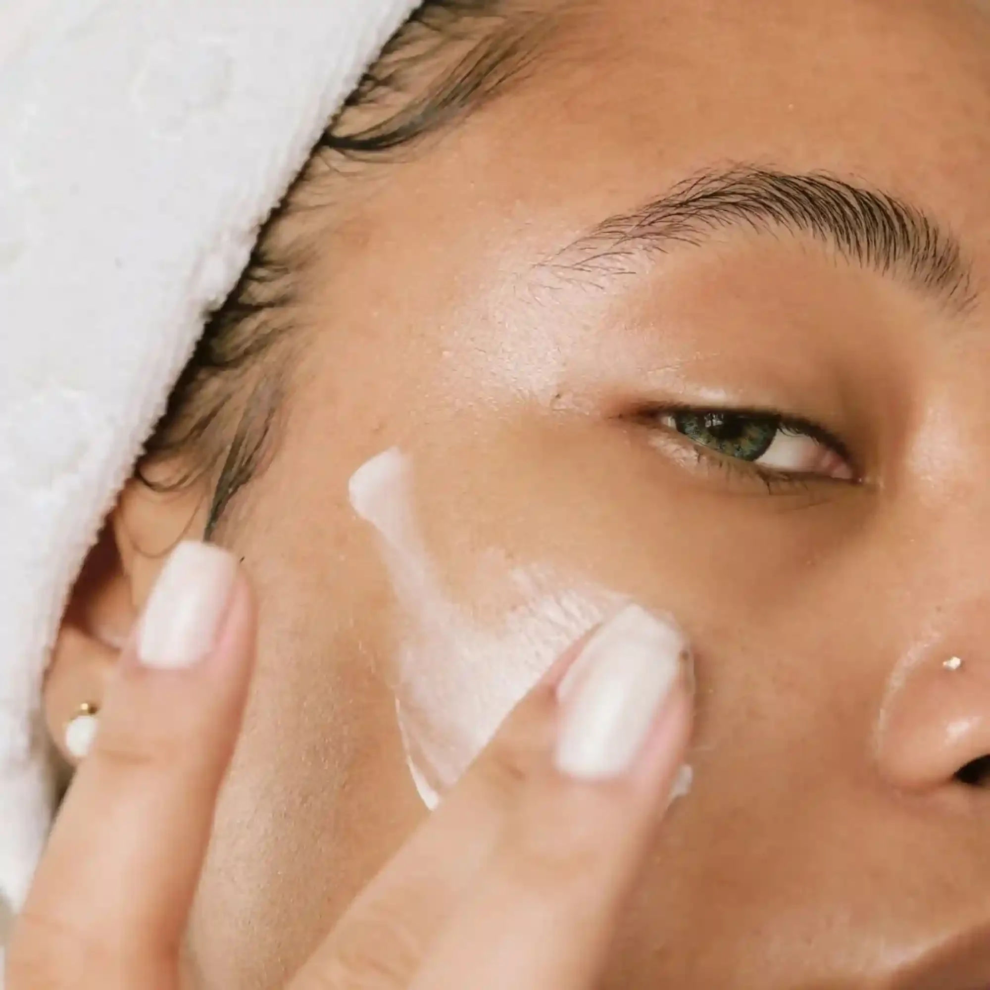 Close-up of a woman applying skincare cream to her face with her finger, with her eye partially closed.