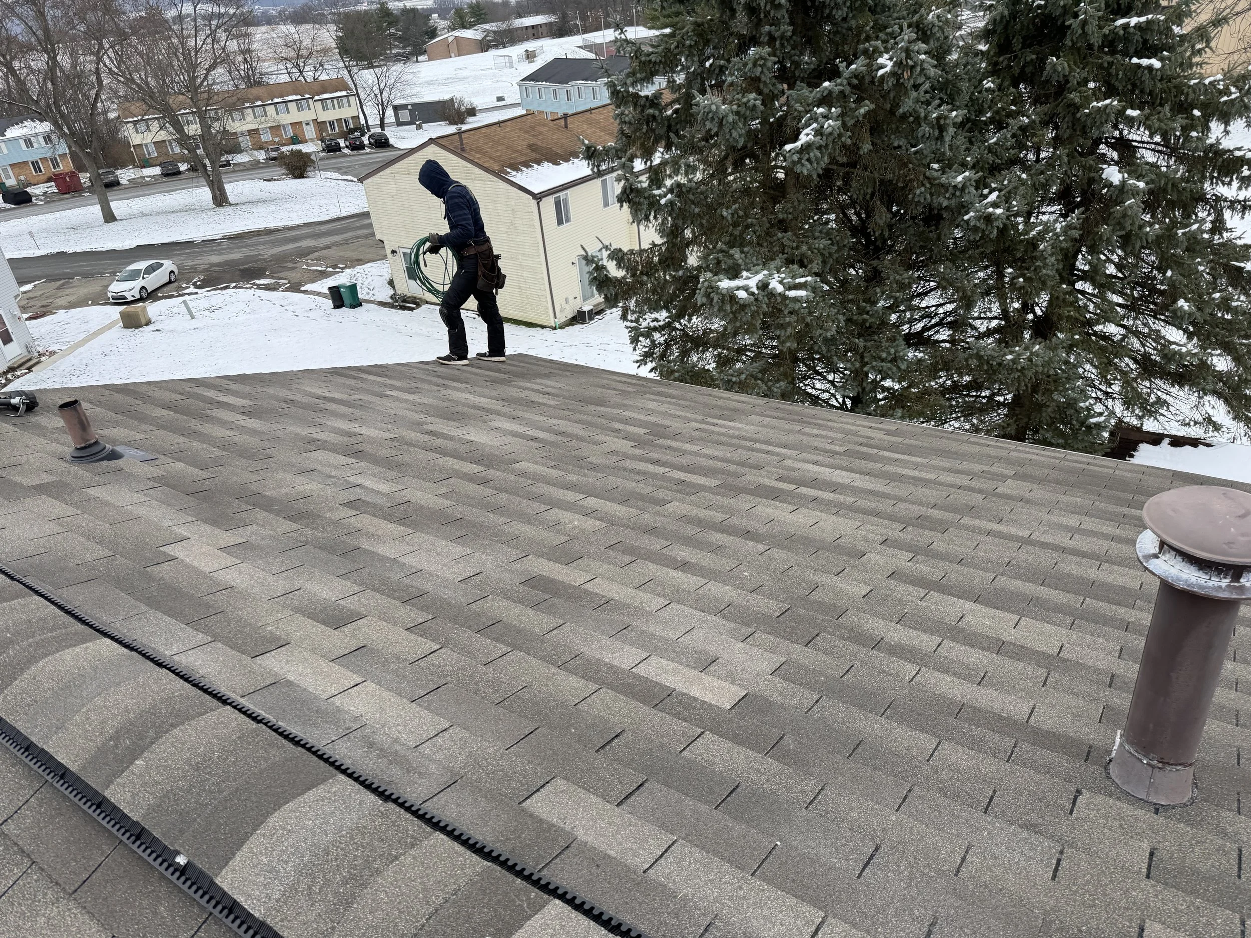 Person in dark hoodie and pants standing on roof, holding a coil of green garden hose, with snow-covered ground, trees, and nearby buildings in background.