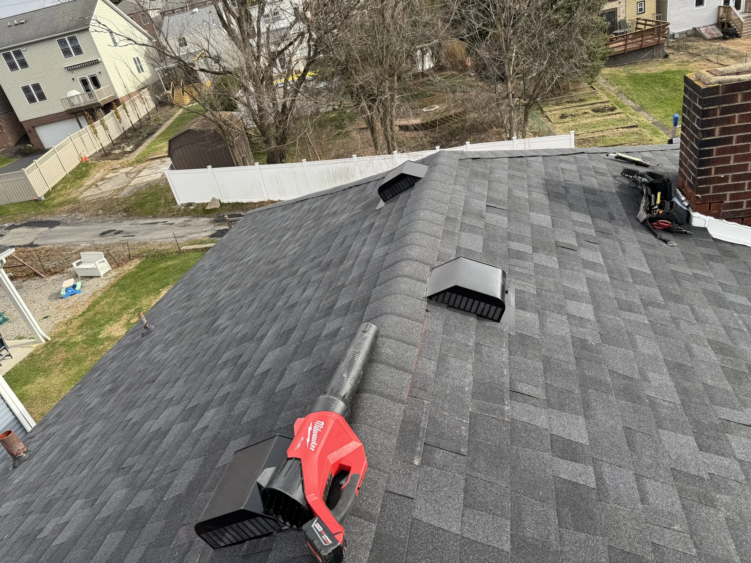 View of a residential roof with a leaf blower and some tools on top, surrounded by neighboring houses and backyards.