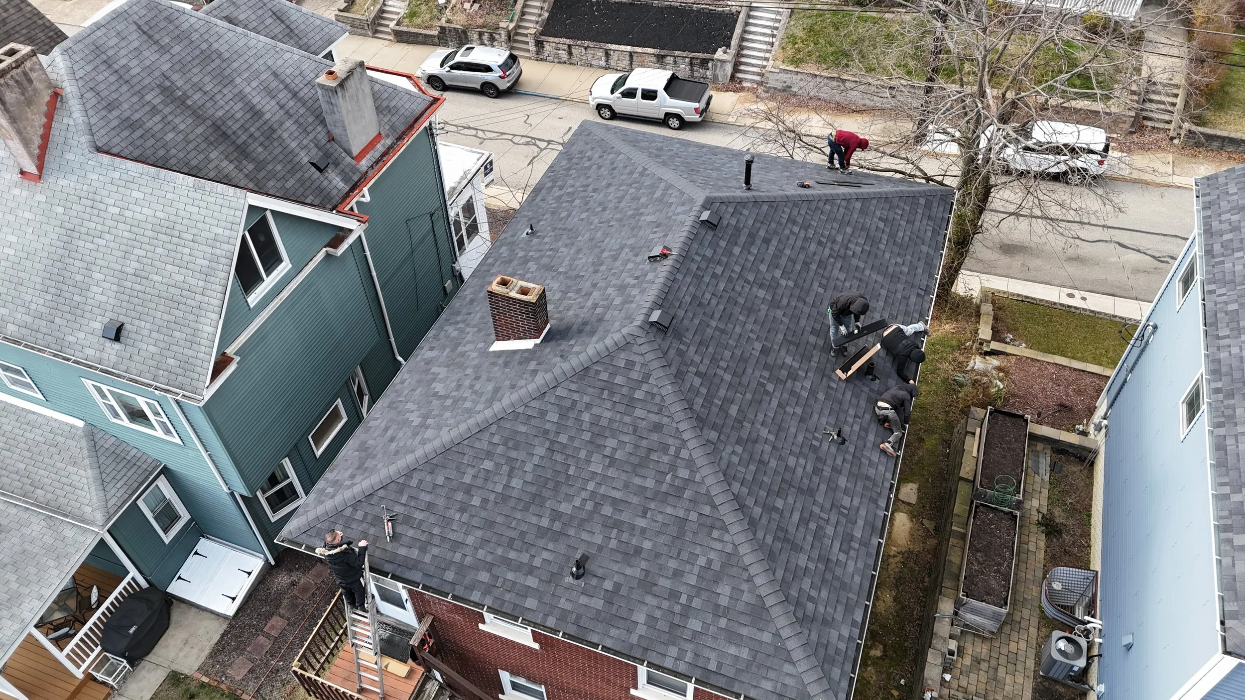 Workers installing or repairing the shingles on a residential roof from a ladder and on the roof itself, with neighboring houses and parked cars visible.