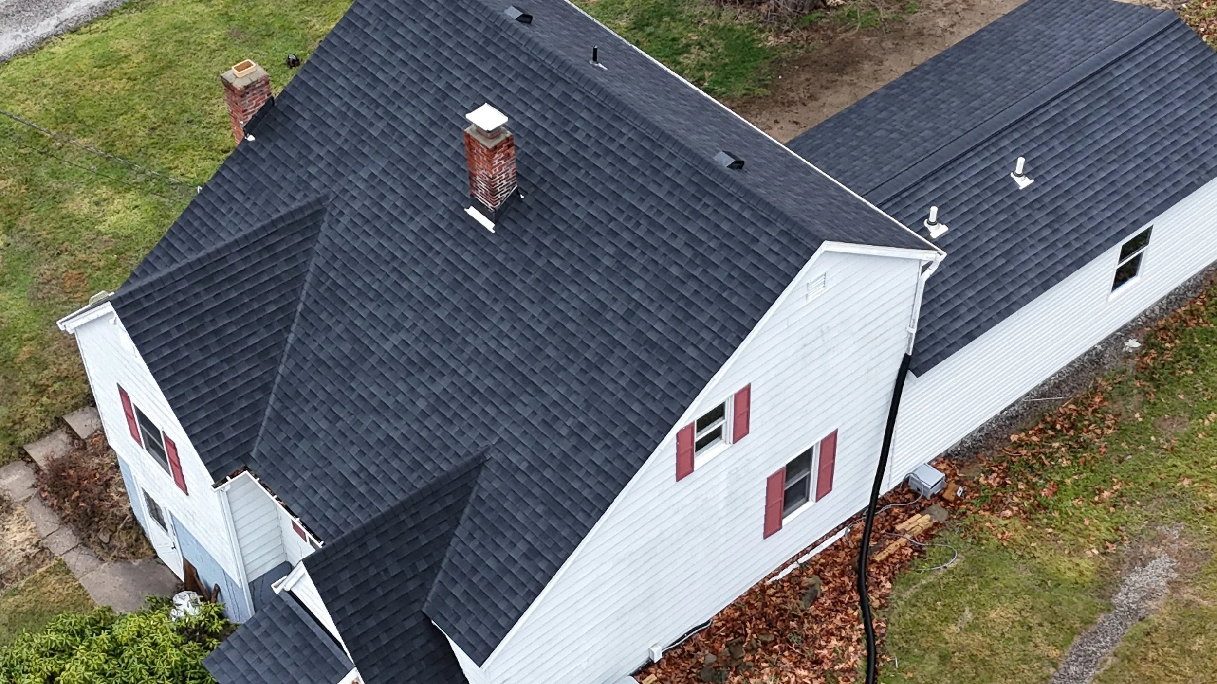 Aerial view of a white house with black roof shingles, red window shutters, and brick chimneys, surrounded by grass, fallen leaves, and a small concrete walkway.