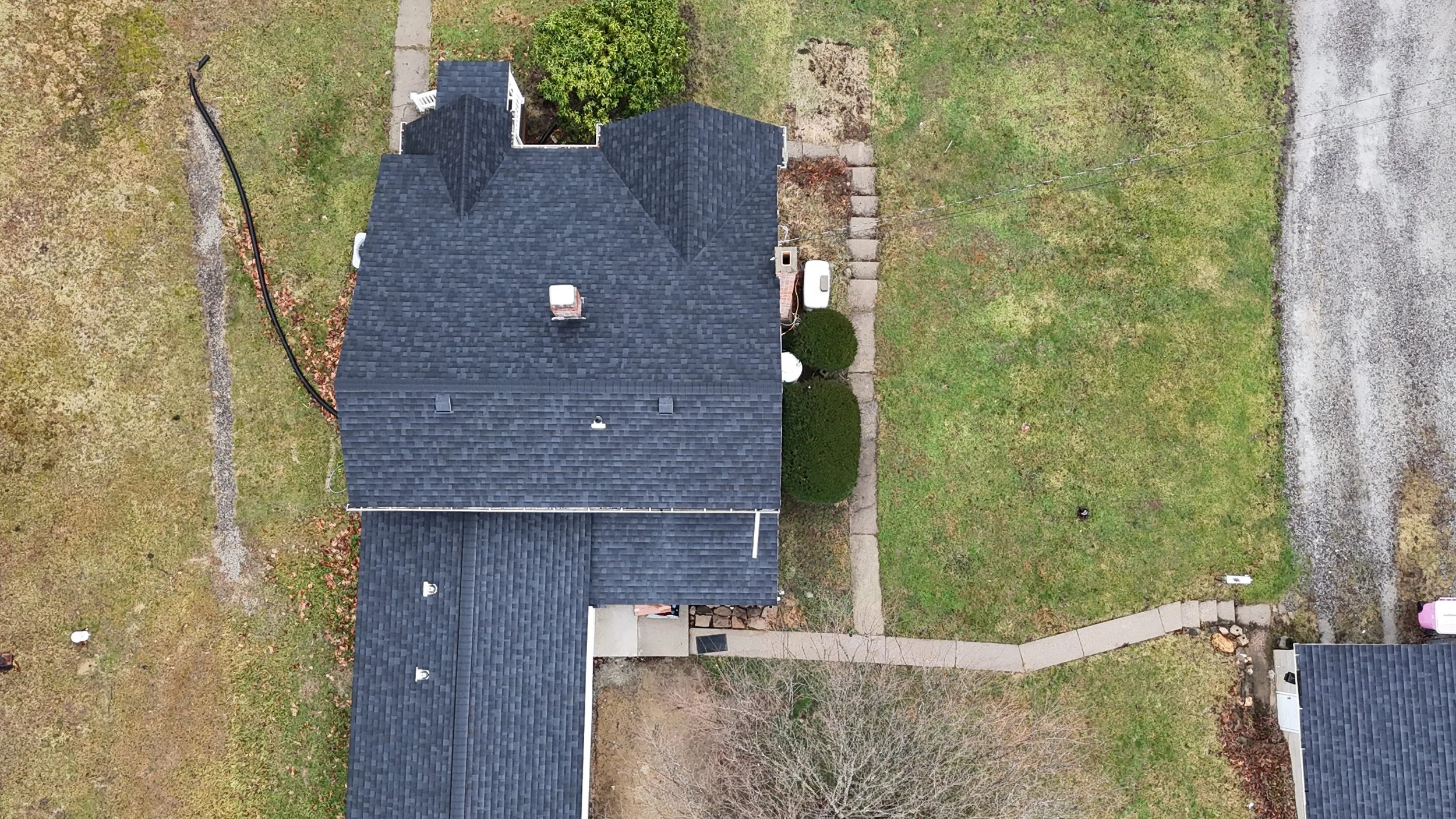 Aerial view of a house with a gray shingle roof, a concrete sidewalk, and a yard with grass and trees.