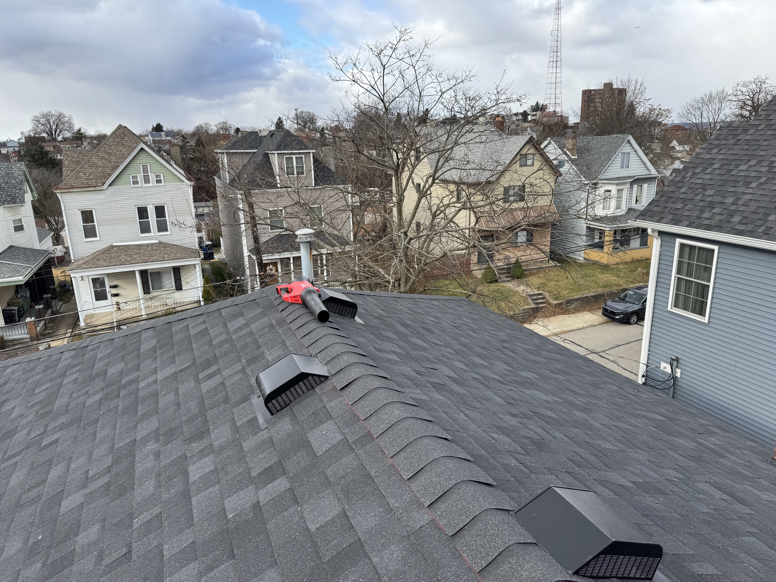 View from a rooftop showing a residential neighborhood with several houses, some trees, and a cloudy sky, with a vent pipe and roof vents visible in the foreground.