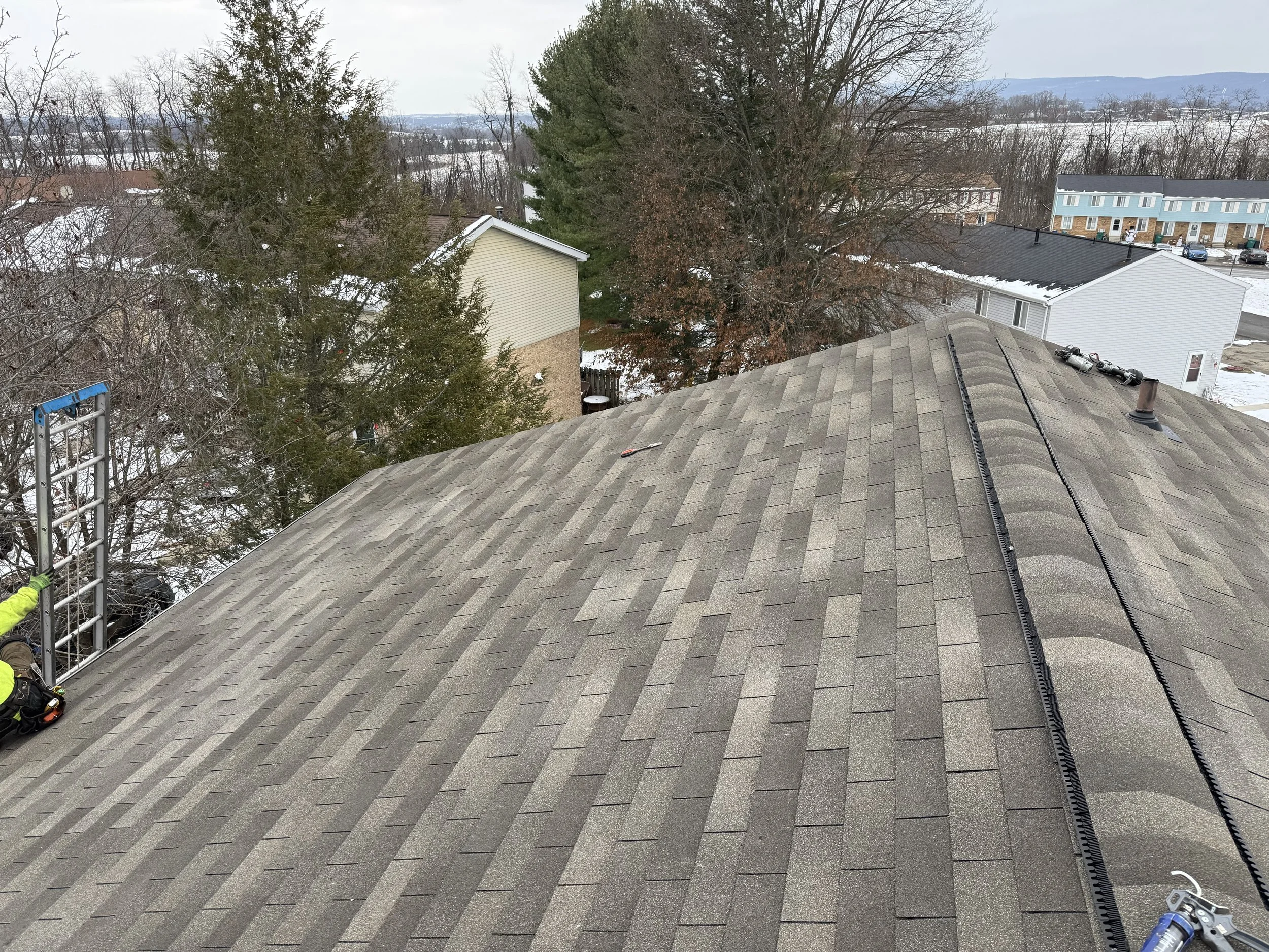 View of a house roof under construction with tools and equipment, surrounded by trees and neighboring houses in a suburban neighborhood.
