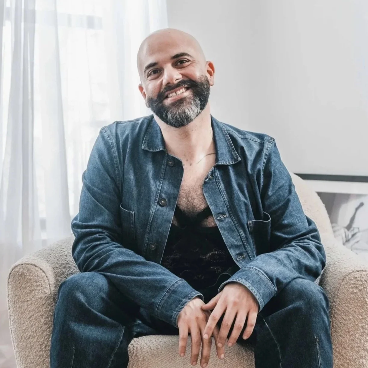 A smiling man with a beard and shaved head sitting on a beige chair in a room with light-colored walls and curtains.