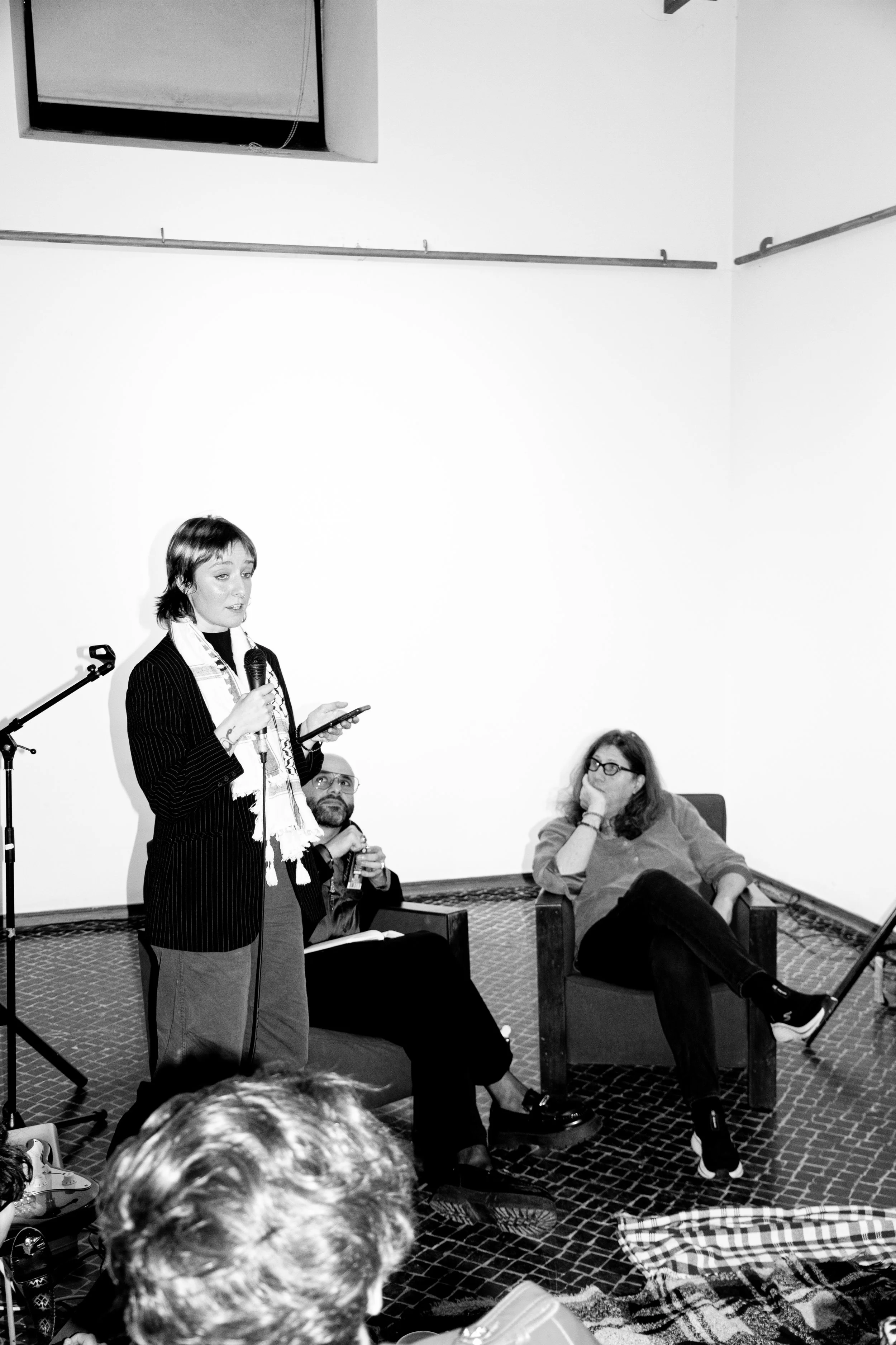 A woman is standing and speaking into a microphone while reading from her phone, with two seated women listening in a room with white walls and a patterned carpet.
