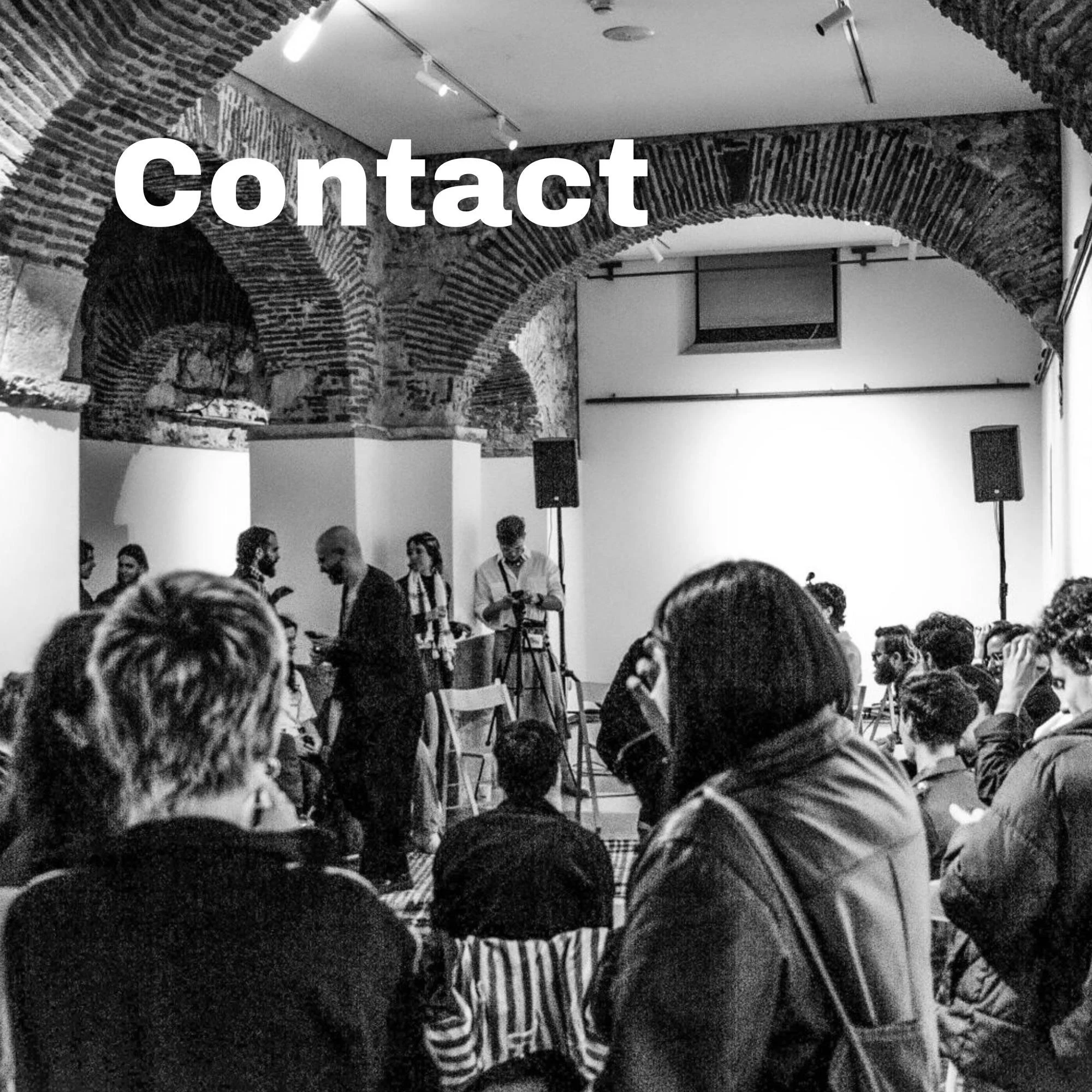A black-and-white photo of an indoor event with exposed brick arches and a seated audience. The word 'Contact' is overlaid across the upper left portion of the image.