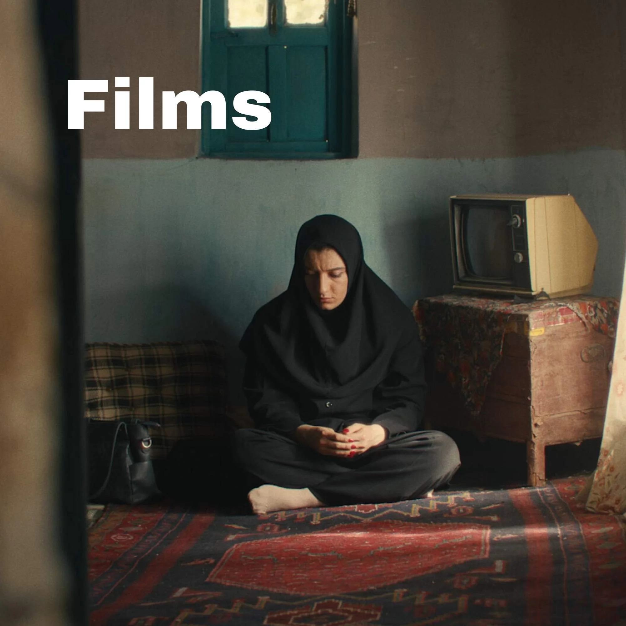 A woman dressed in black traditional attire sitting on a patterned rug in a small, modest room with a blue door, an old television on a wooden chest, and textured walls.