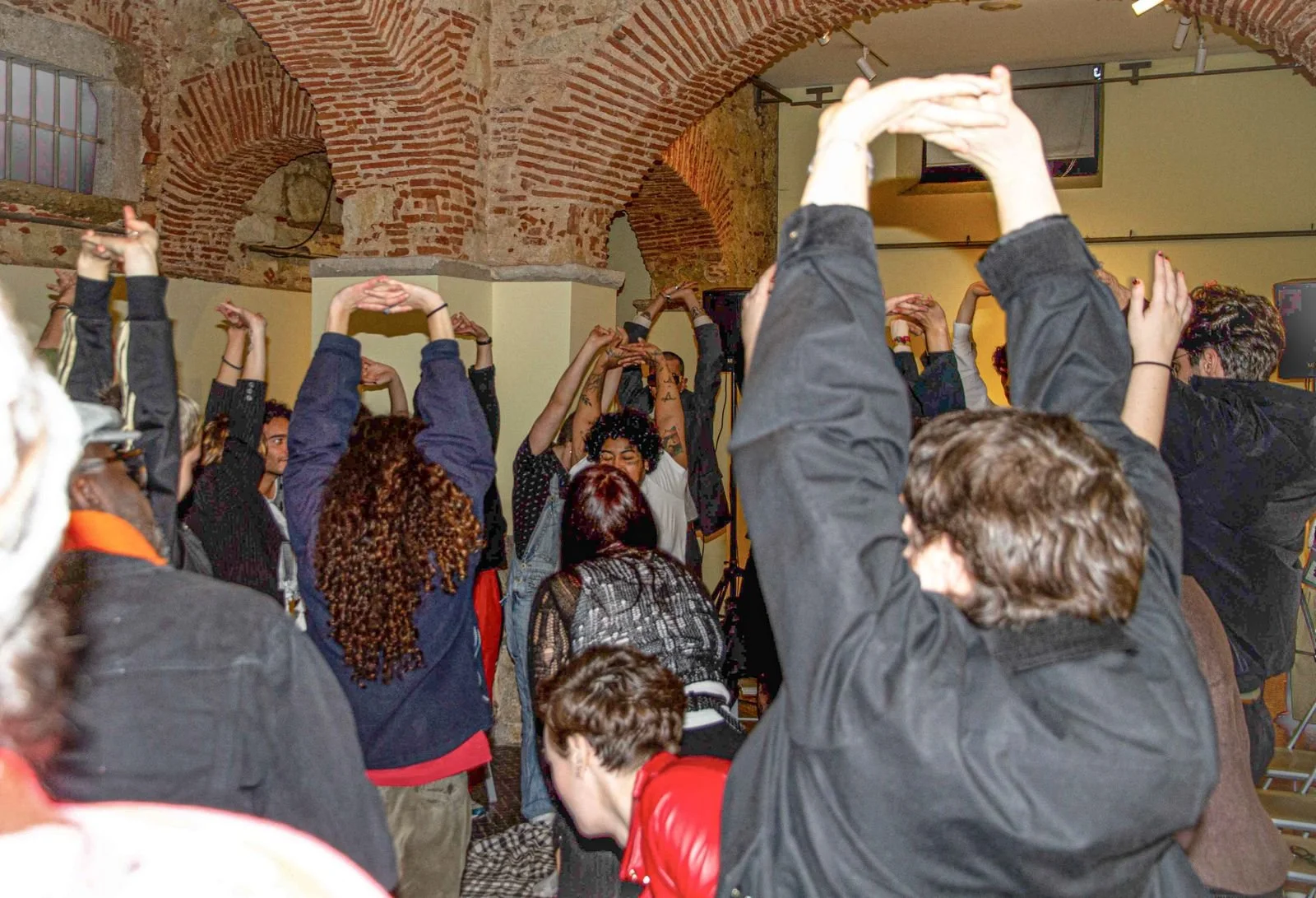 Group of people raising their hands in a room with brick arches and yellow walls, participating in a dance or exercise activity.
