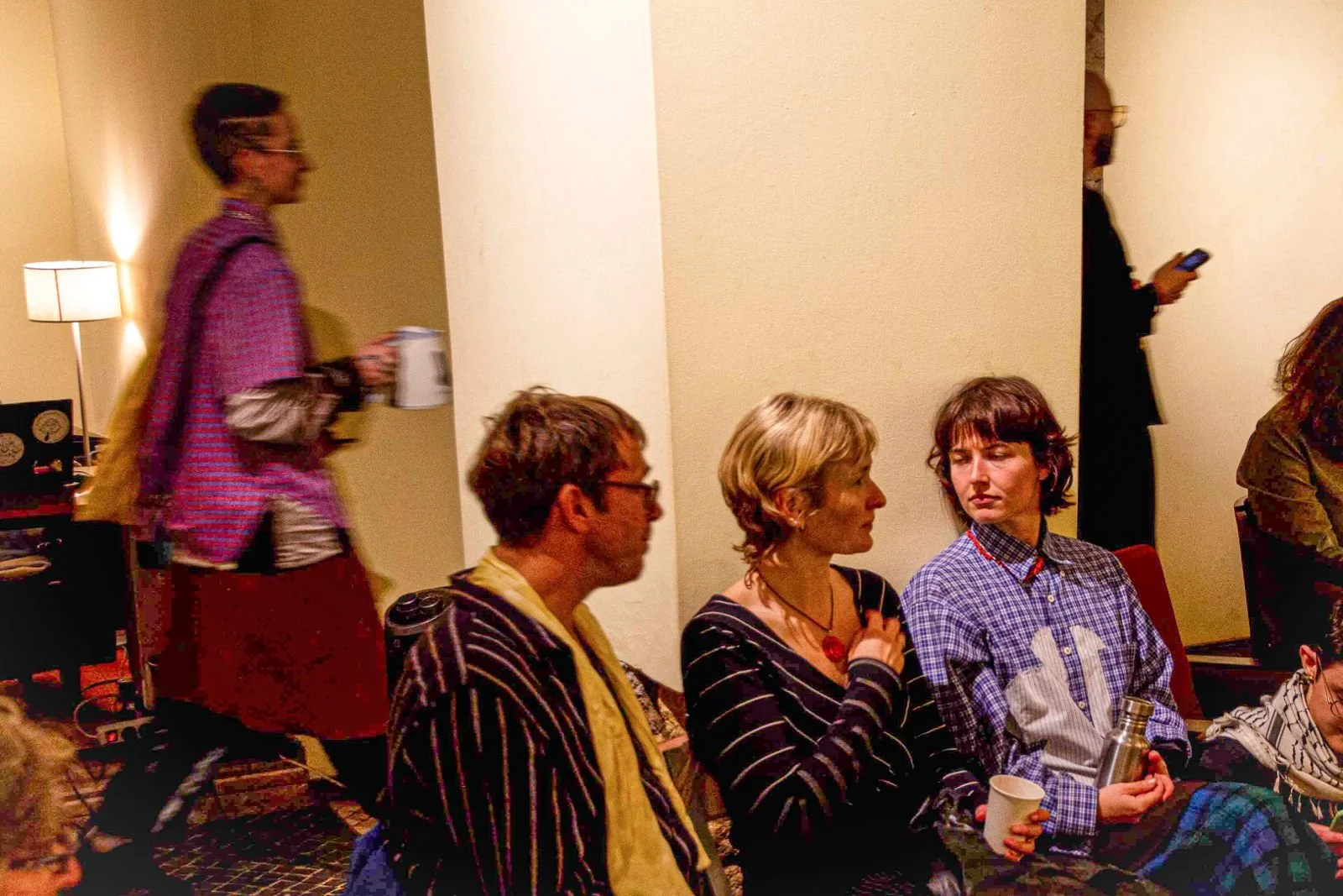 Group of people sitting and talking in a room with in a casual setting; one woman holding a mug, another person holding a water bottle, others with cups, in a cozy environment with warm lighting.