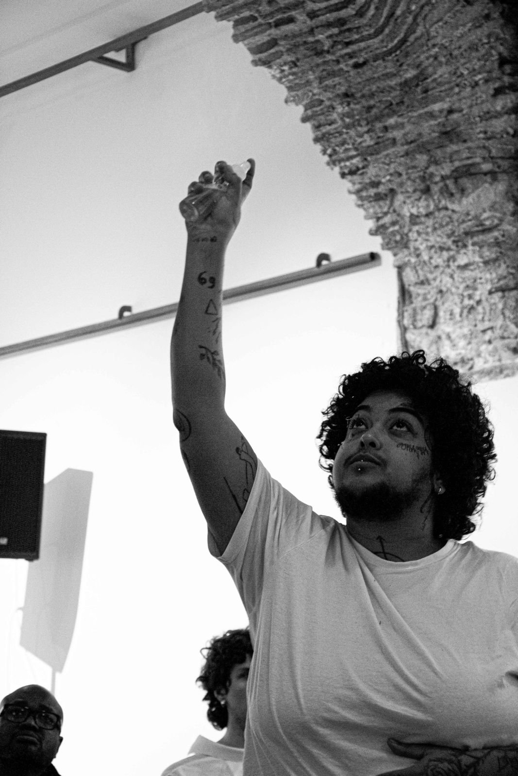 Luan Okun with curly hair and tattoos on their arms raising a glass in a toast in an indoor setting with a brick wall and ceiling beam visible at JAHA Film Festival.