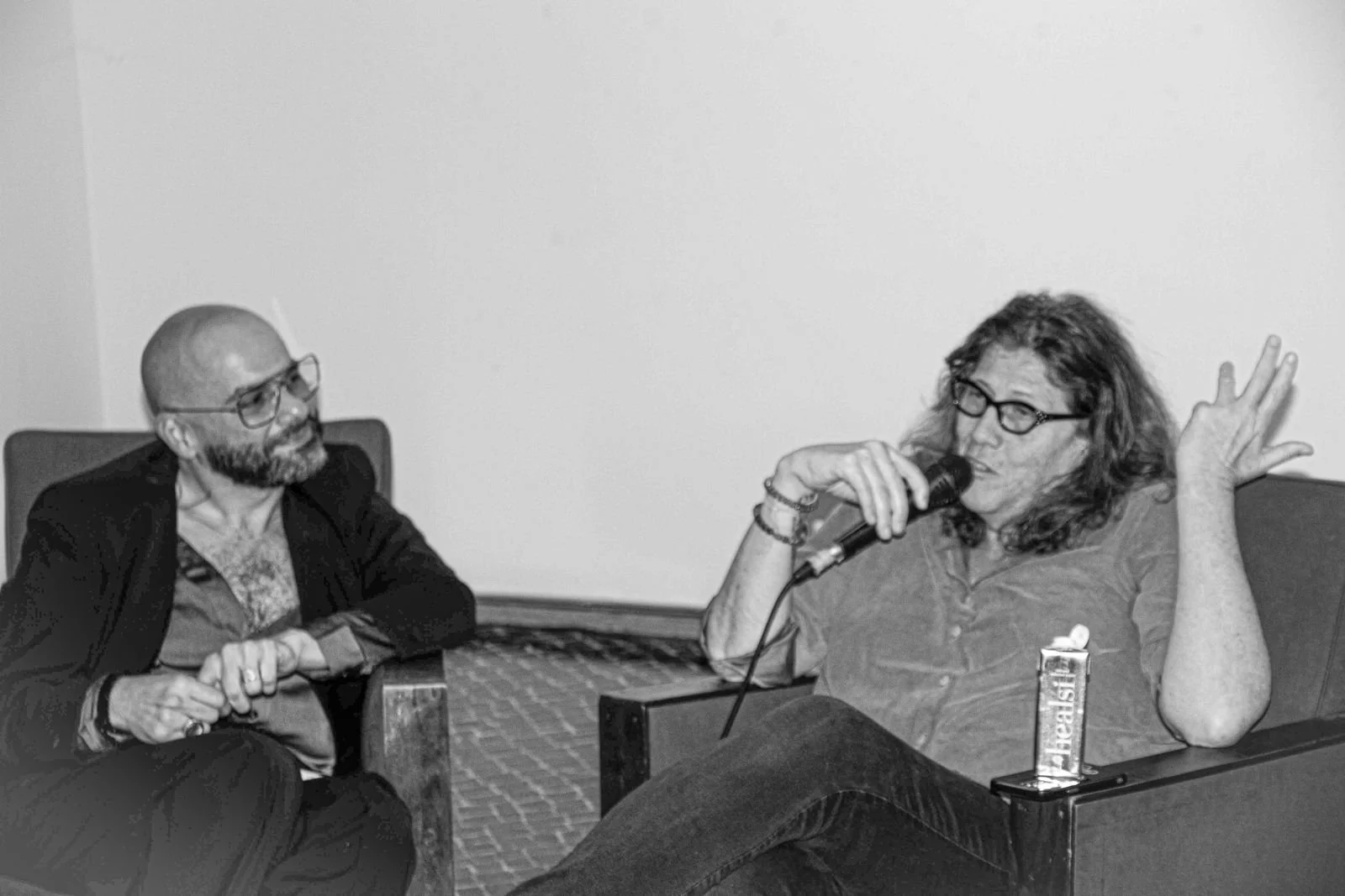 Susan Stryker and Mama Ganuush sitting on chairs having a conversation; one person is holding a microphone and gesturing "peace" with their fingers; the other person is listening attentively. Jaha Film Festival closing gala.