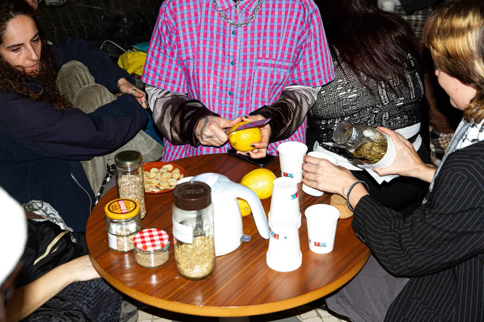 People gathered around a table having snacks and pouring beverages, with jars of oats, a lemon, and disposable cups on the table.