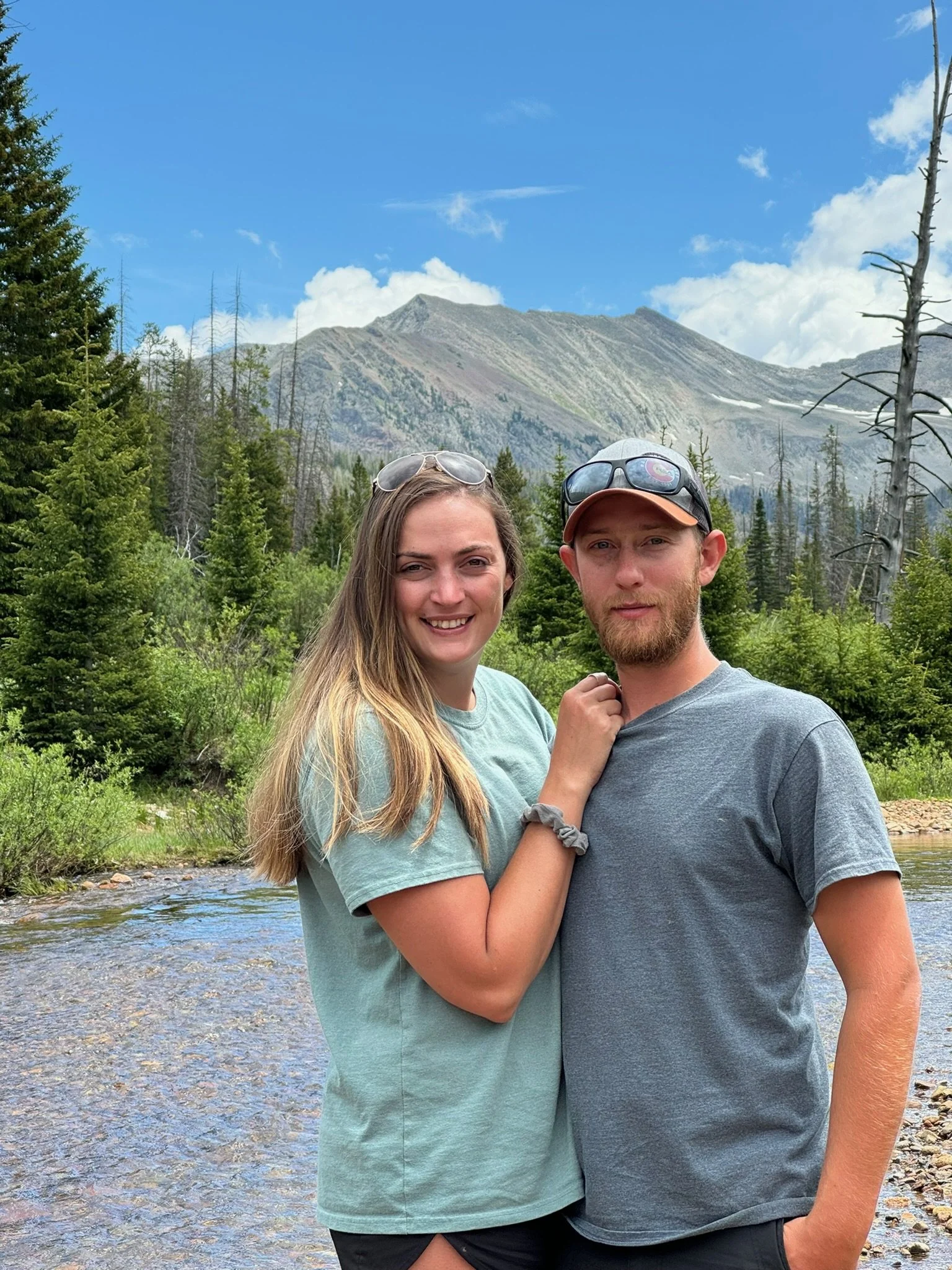 A smiling young woman and a man stand close together in an outdoor setting with a mountain, trees, and a river in the background, on a partly cloudy day. Owner Shawn and his girlfriend