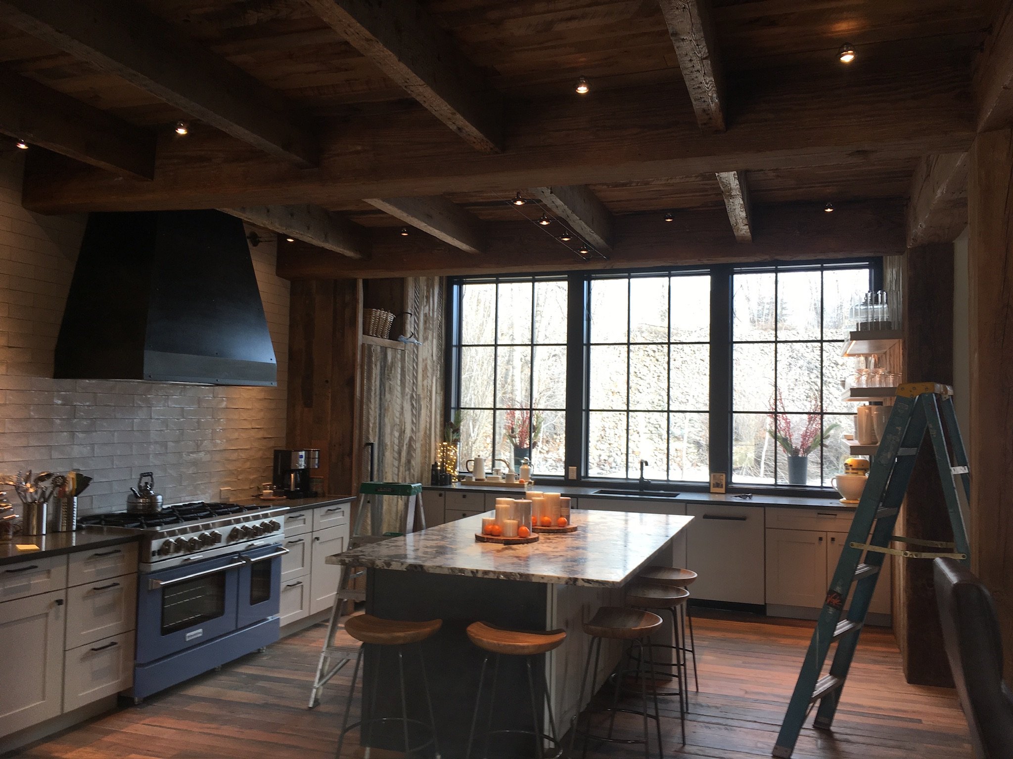 Modern kitchen with wooden beams on the ceiling, large windows, a marble island, white cabinets, a stove, and decorative candles on the island. Lighting installed on the ceiling and cabinets