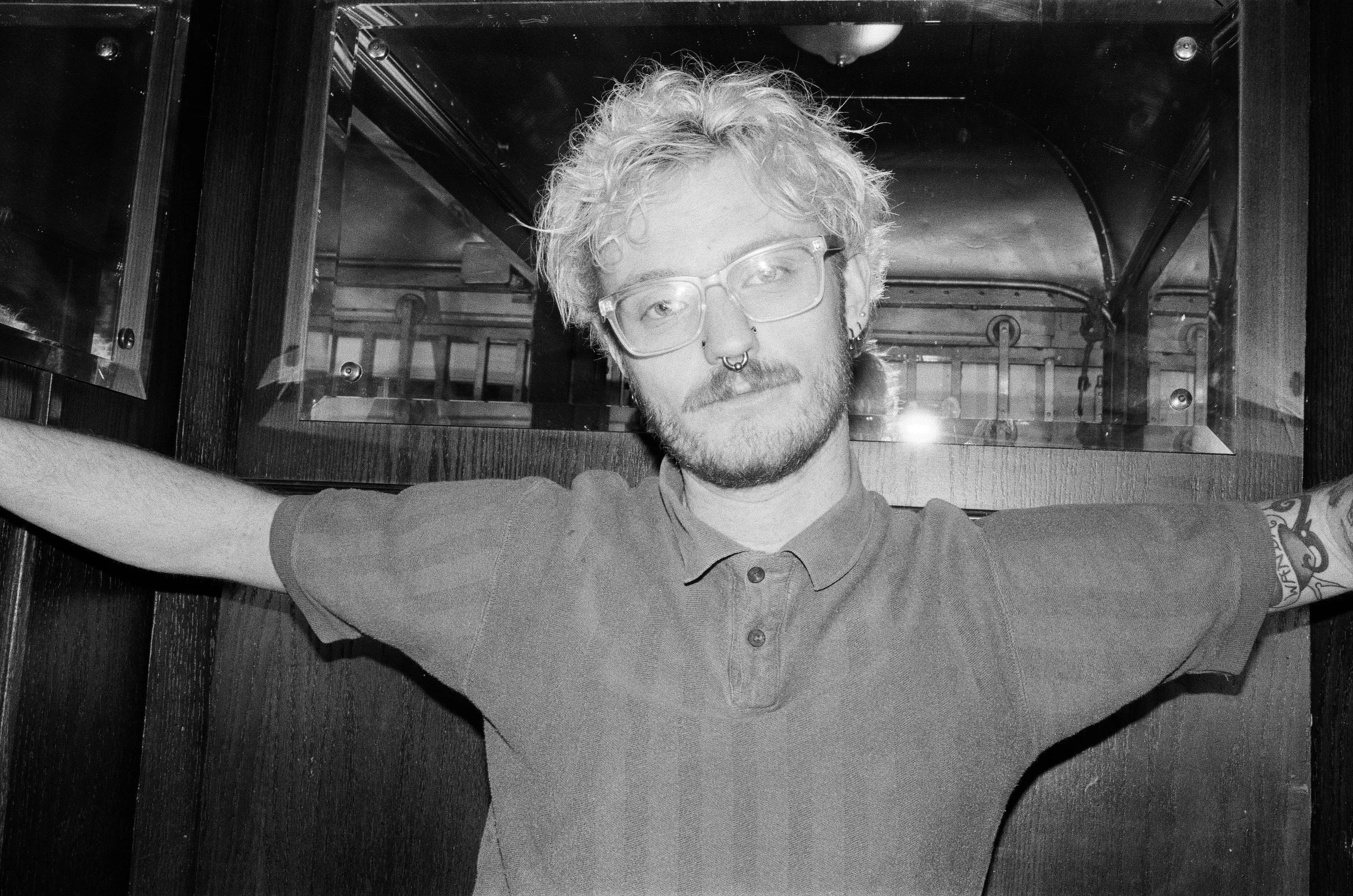 A young man with glasses, a nose ring, and tattoos on his arms, wearing a polo shirt, is taking a selfie with his arms outstretched. He is standing in a room with wooden walls and a large mirror or glass behind him. The photo is in black and white.