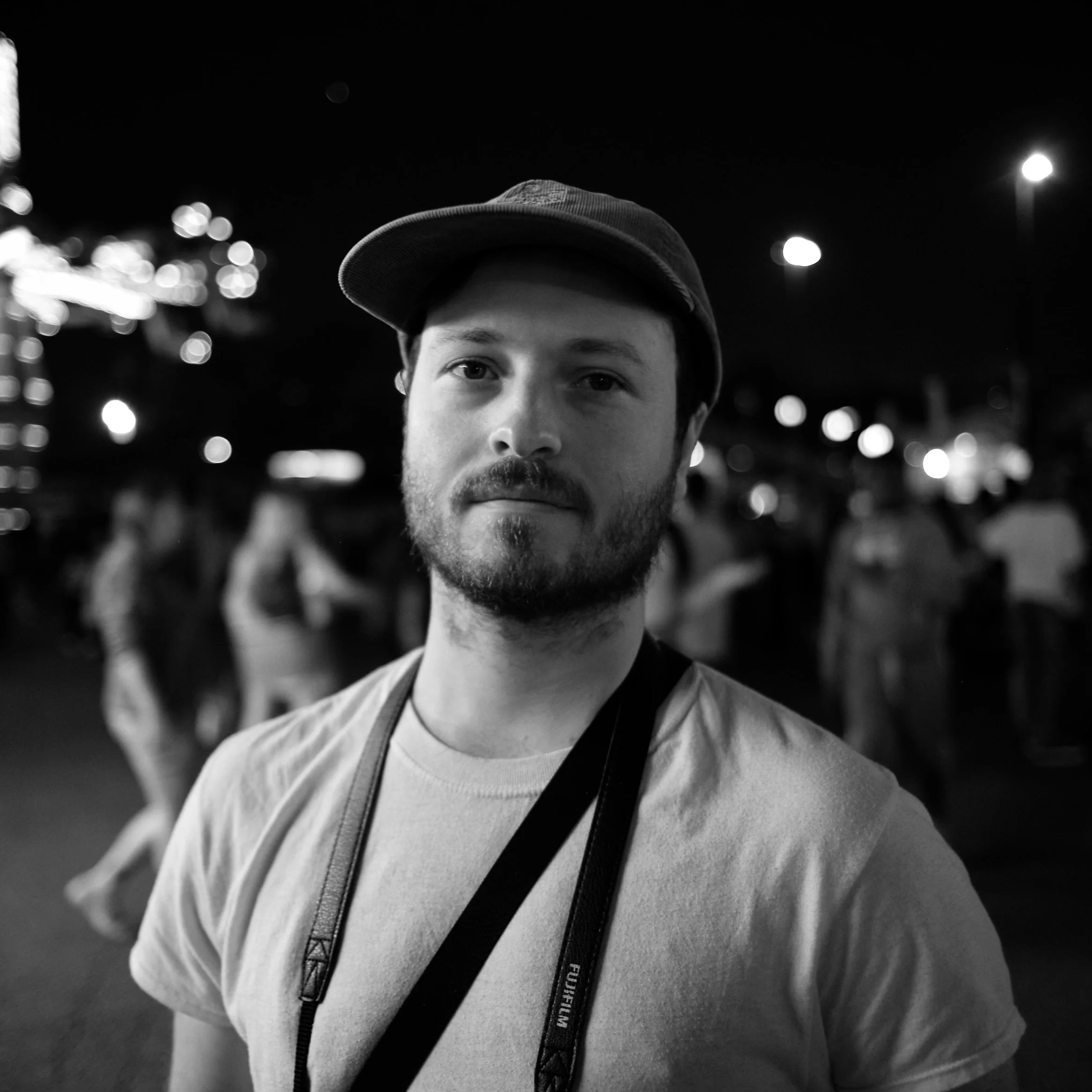 Close-up of a young man wearing a baseball cap and a backpack, standing outdoors at night with blurred city lights in the background.