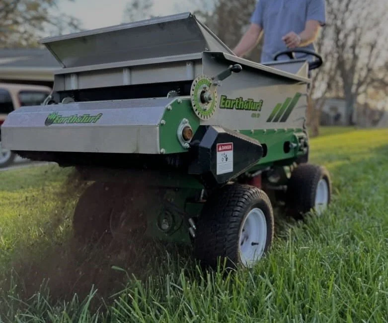 A person operating an Earthstaur gauge in a grassy field, with a tractor in the background.