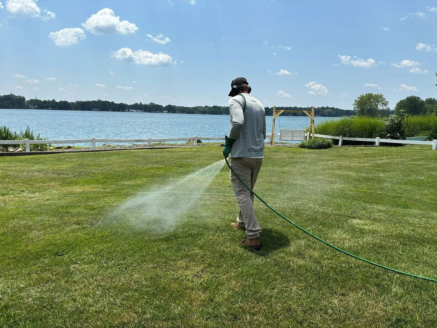 A person wearing a gray long-sleeve shirt, tan pants, and a black cap is watering the grass with a garden hose near a lake on a sunny day with blue skies and a few white clouds.