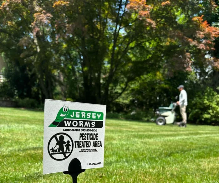Sign indicating Jersey worms landscaping pesticide-treated area, with a person using a lawn mower in the background in a grassy yard with trees.