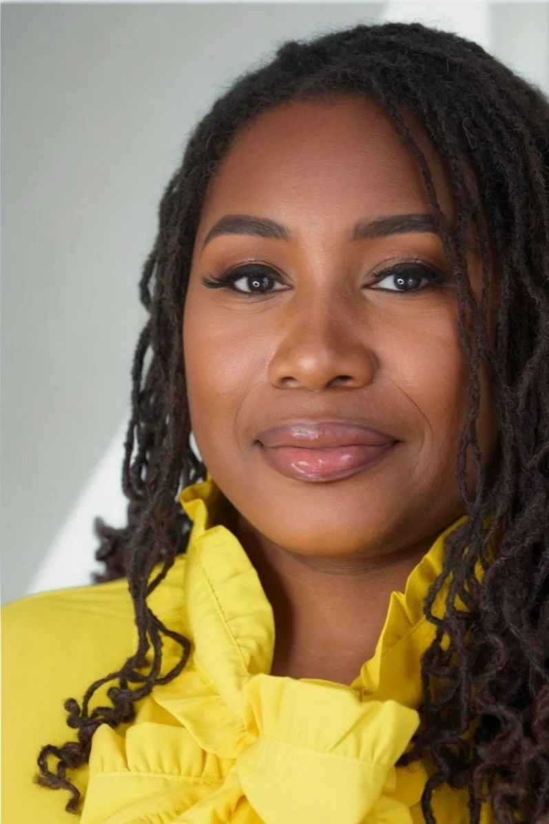 A woman with dark curly hair, wearing a bright yellow ruffled blouse, is looking at the camera with a soft smile.