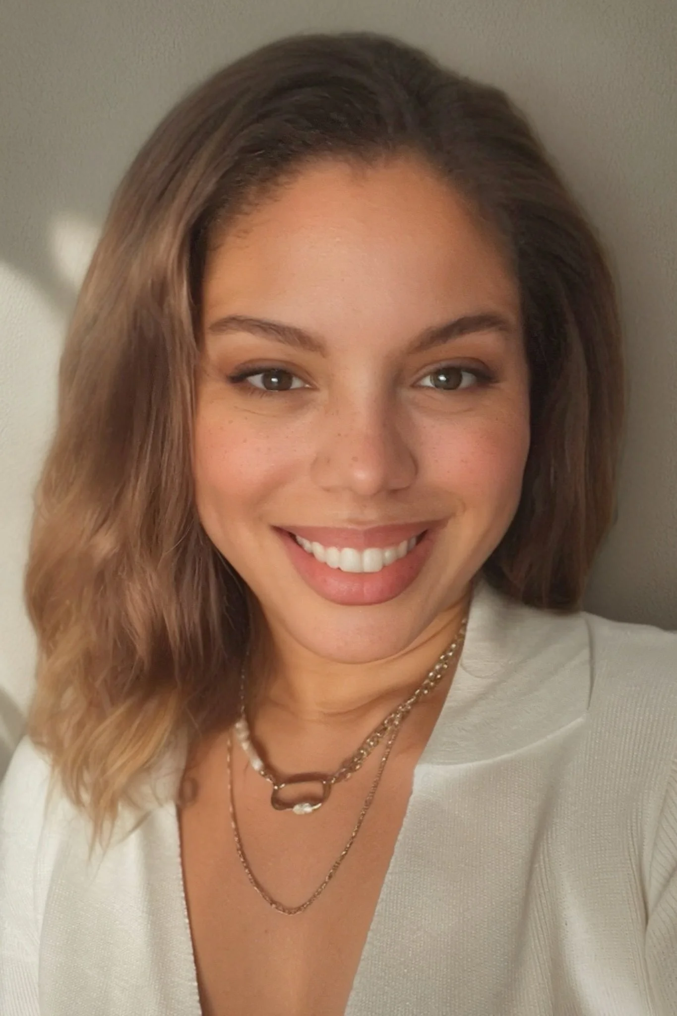 A woman with shoulder-length wavy hair smiling, wearing neutral makeup, a white top, and layered necklaces.