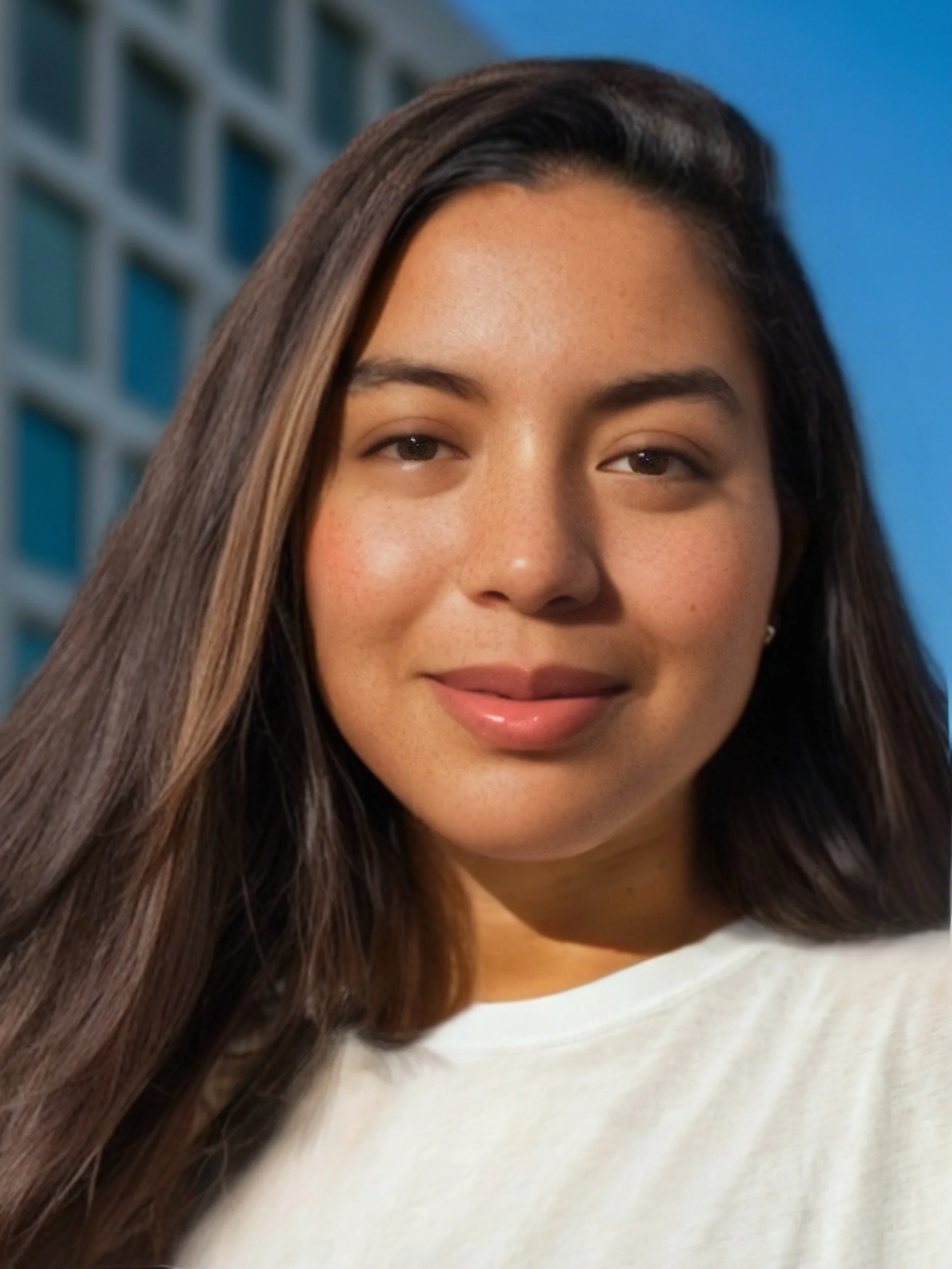Close-up of a young woman with long dark hair smiling, with a modern building and blue sky in the background.