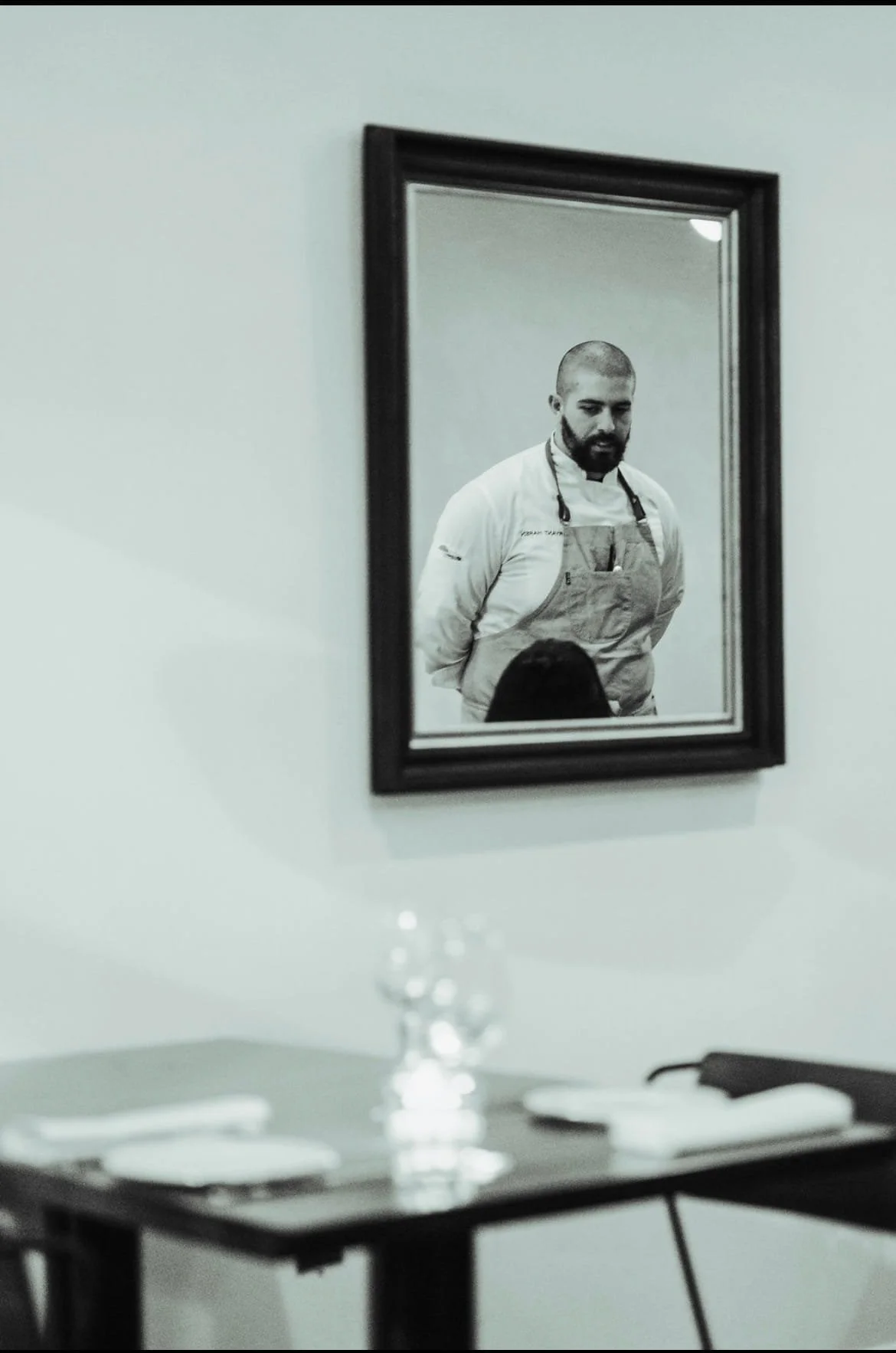 A man in a chef's jacket and apron is looking at his reflection in a mirror on a white wall.