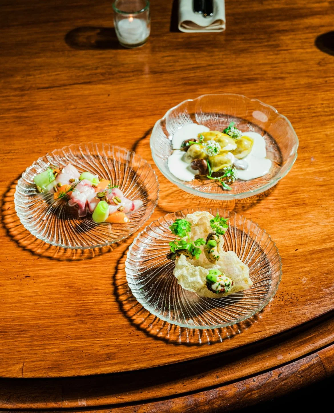 Three glass plates with gourmet appetizers on a wooden table, featuring colorful ingredients, herbs, and garnishes.
