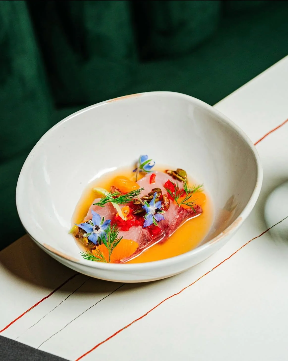 A plated dish featuring raw fish, garnished with edible flowers, herbs, and a flavorful broth.