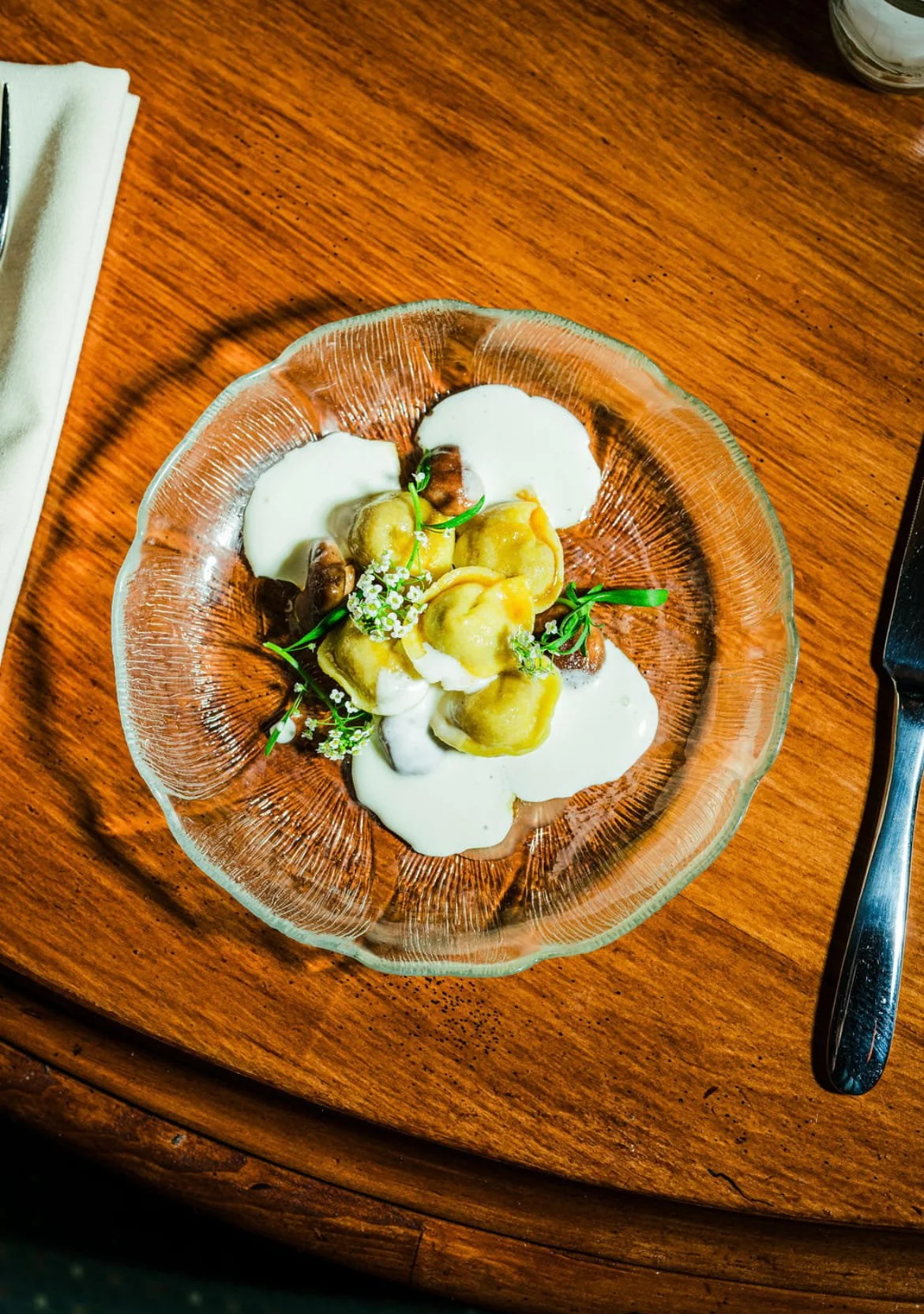 A glass bowl containing tortellini pasta with cream sauce and garnished with microgreens and edible flowers on a wooden table.