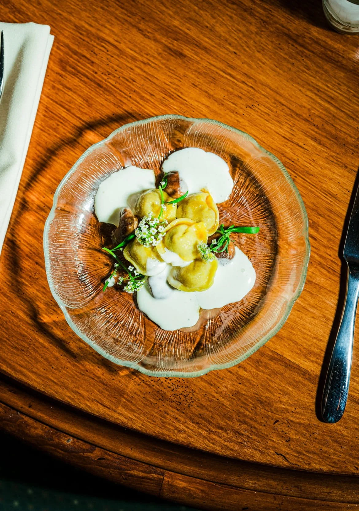 A glass bowl of ravioli pasta with white creamy sauce, garnished with herbs and tiny white flowers, on a wooden table.