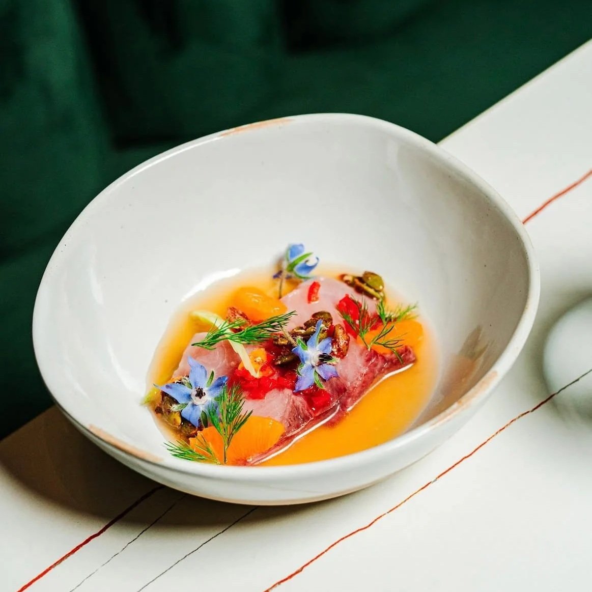 A white bowl with a dish garnished with edible flowers, herbs, and sliced vegetables over a runny yolk and slices of raw fish or meat.