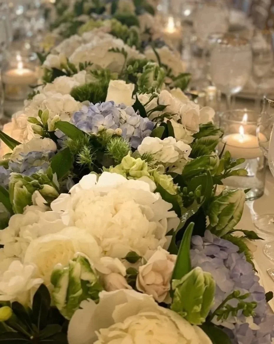 Close-up of a long floral centerpiece with white, green, and light purple flowers, surrounded by glass candle holders with lit candles on a dining table.