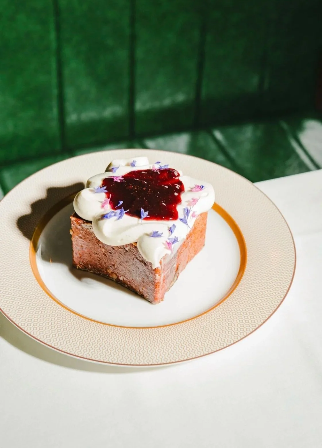 A square slice of chocolate cake with cream and purple flower petals, topped with berry jam, on a decorative beige plate.