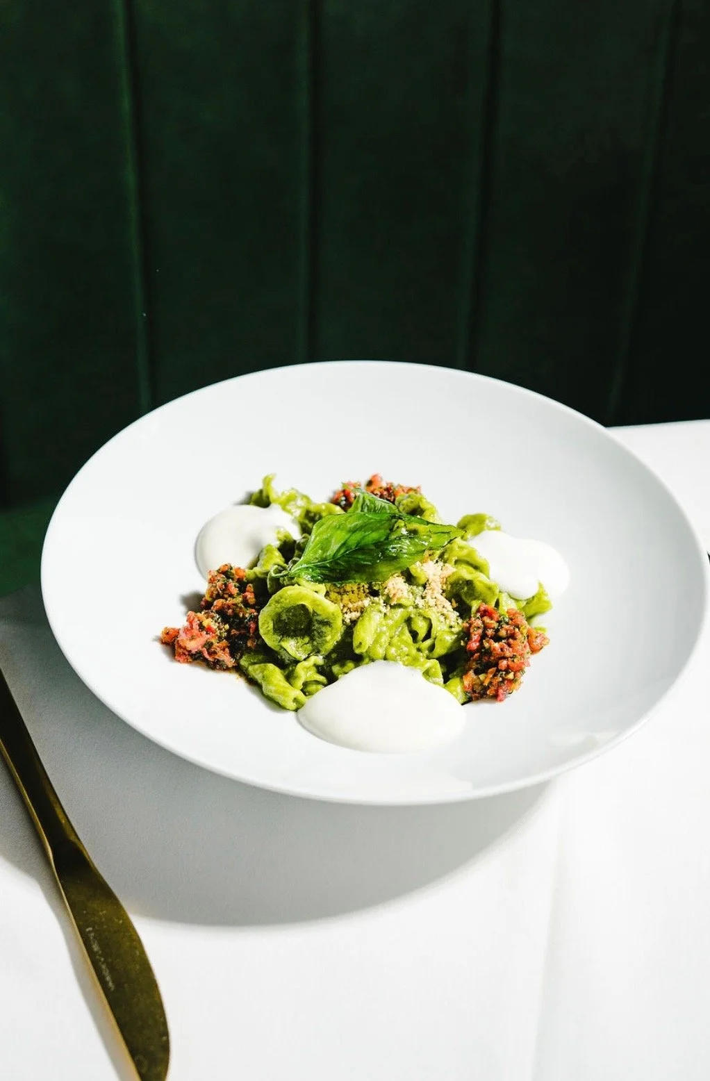 A white bowl of pasta with green sauce, garnished with fresh basil, accompanied by dollops of white sauce, on a white table with a gold knife nearby.