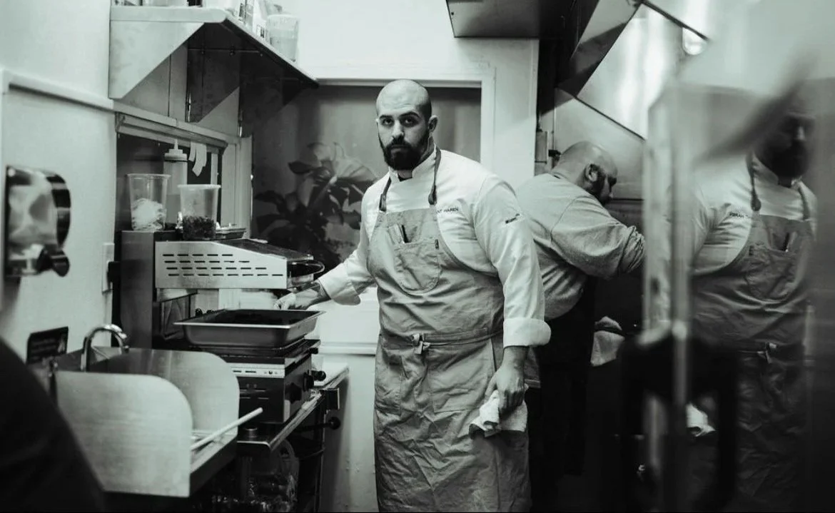 A chef with a beard and a bald head stands in a kitchen, wearing a white chef's coat and an apron, looking at the camera. Two other chefs are working behind him.