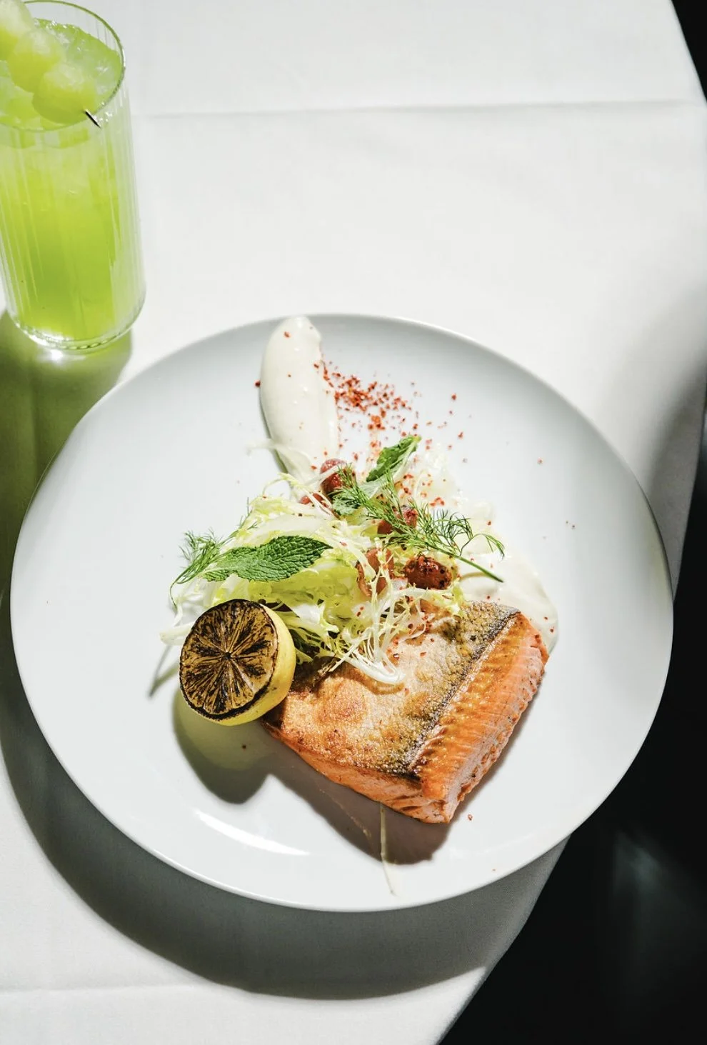 A plate with a cooked salmon fillet, garnished with fresh herbs, a lemon slice, and served with a side of salad and a dollop of sauce. A glass of green juice is visible on the side.
